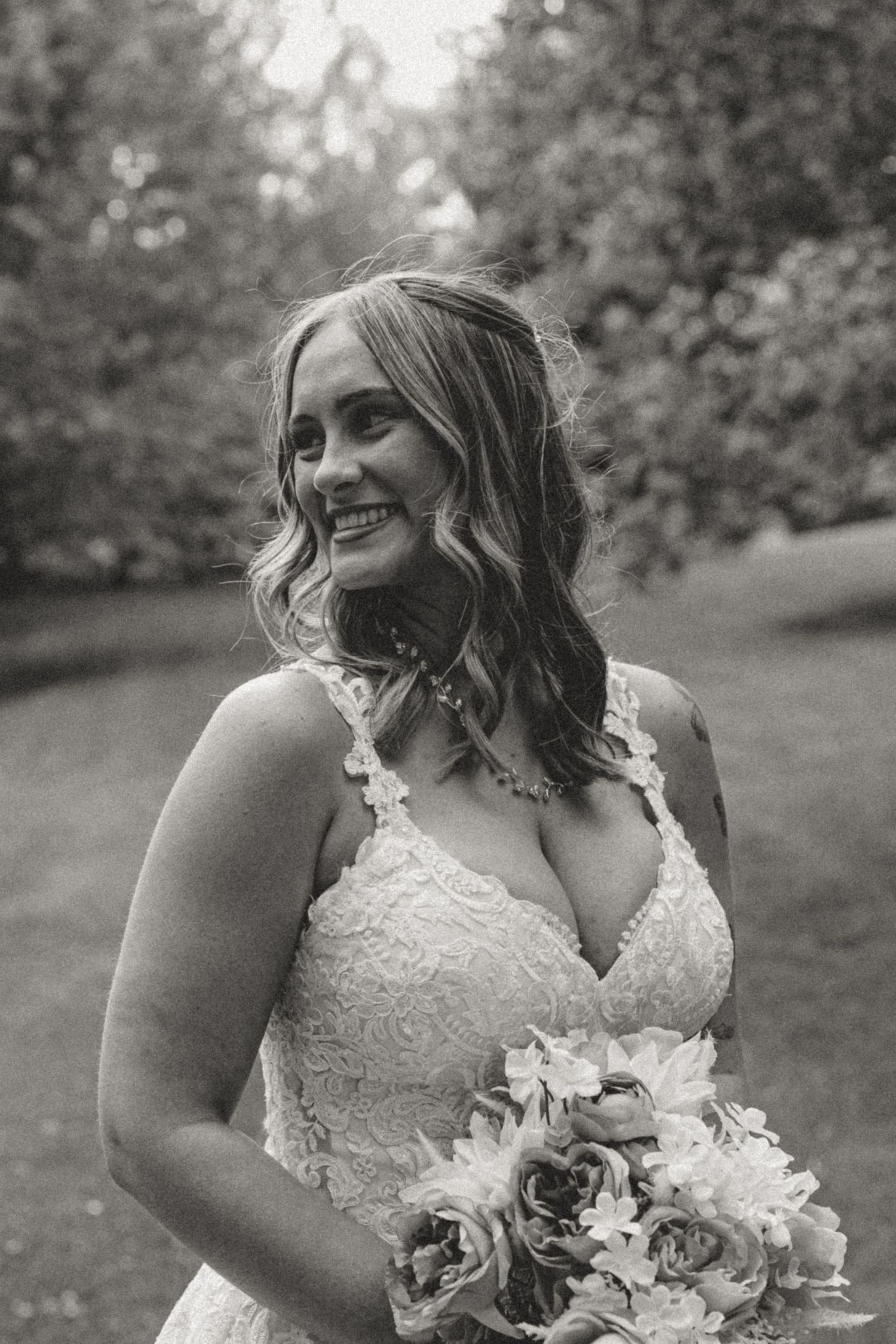 A smiling bride in a lace wedding dress holds a bouquet and looks to the side in a black and white outdoor portrait.