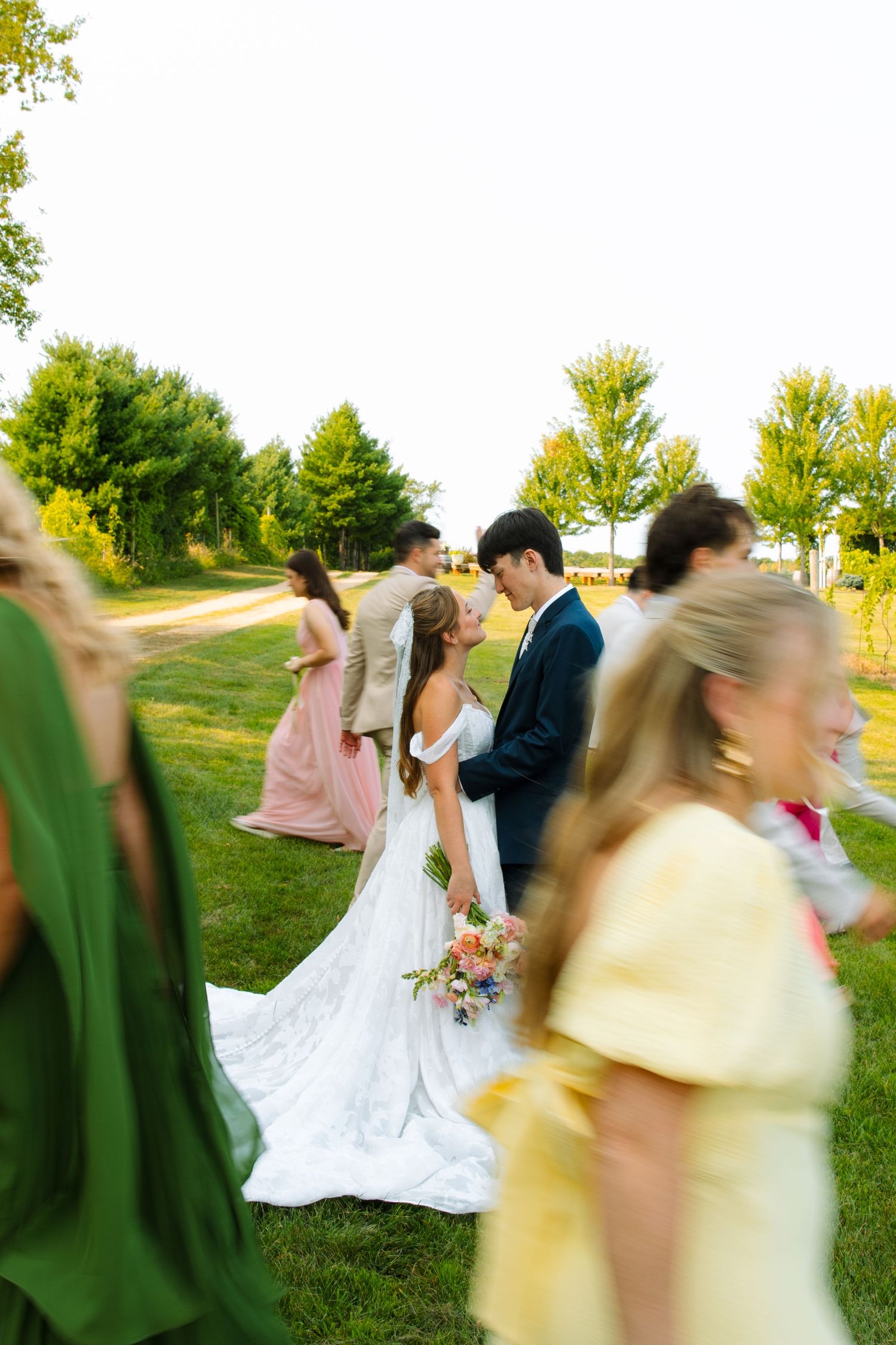 A motion-blurred wedding party in colorful dresses and suits walks across a sunny lawn around a bride and groom in an embrace.