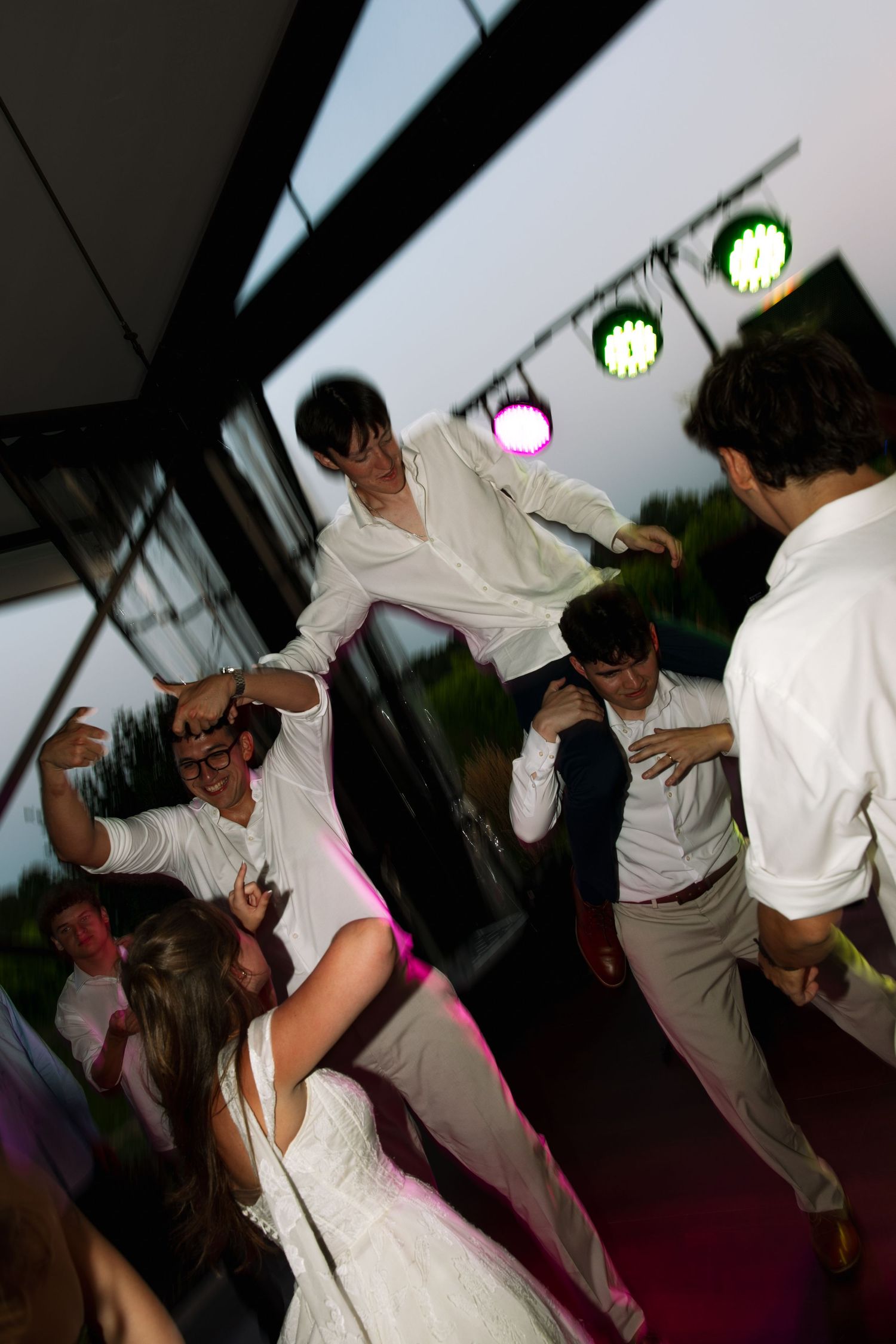 Wedding guests in white shirts dance and lift a man into the air under colorful stage lights at an evening outdoor reception.