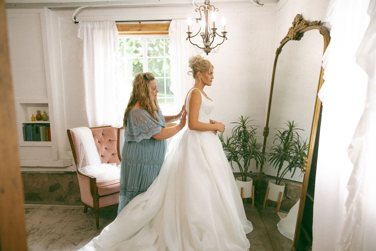 A bridesmaid helps a bride in a flowing white wedding gown get ready beside an antique mirror in a bright room.