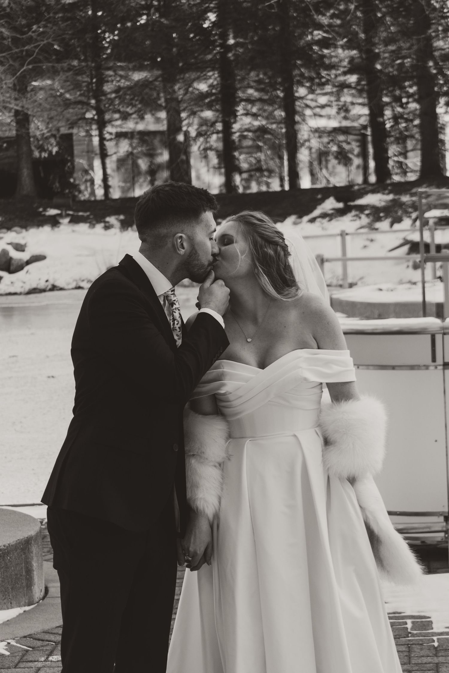 Black and white photo of a groom kissing his bride in an off-shoulder satin gown and fur wrap beside a snowy winter landscape.