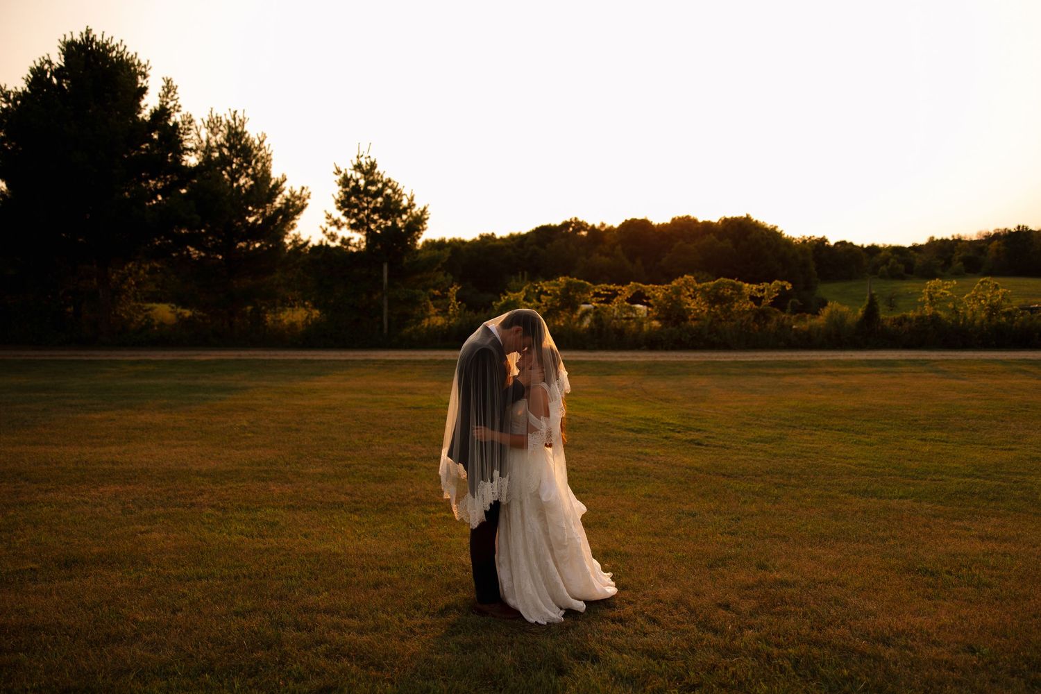 Bride and groom embrace under a long veil in an open field at golden hour, with trees and vineyards in the background.