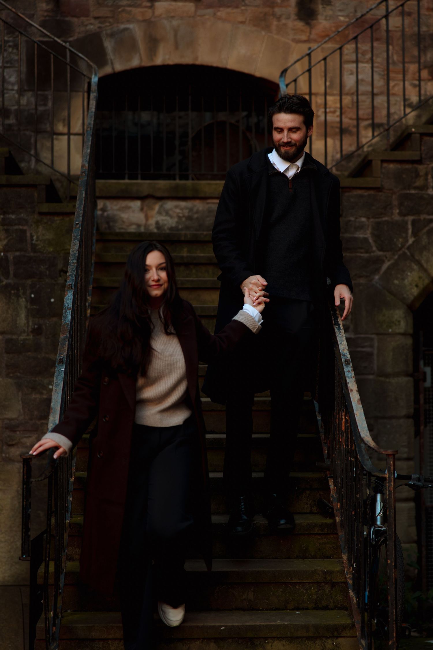 Couple in dark coats holding hands while walking down worn stone steps with iron railings during an engagement photo session.