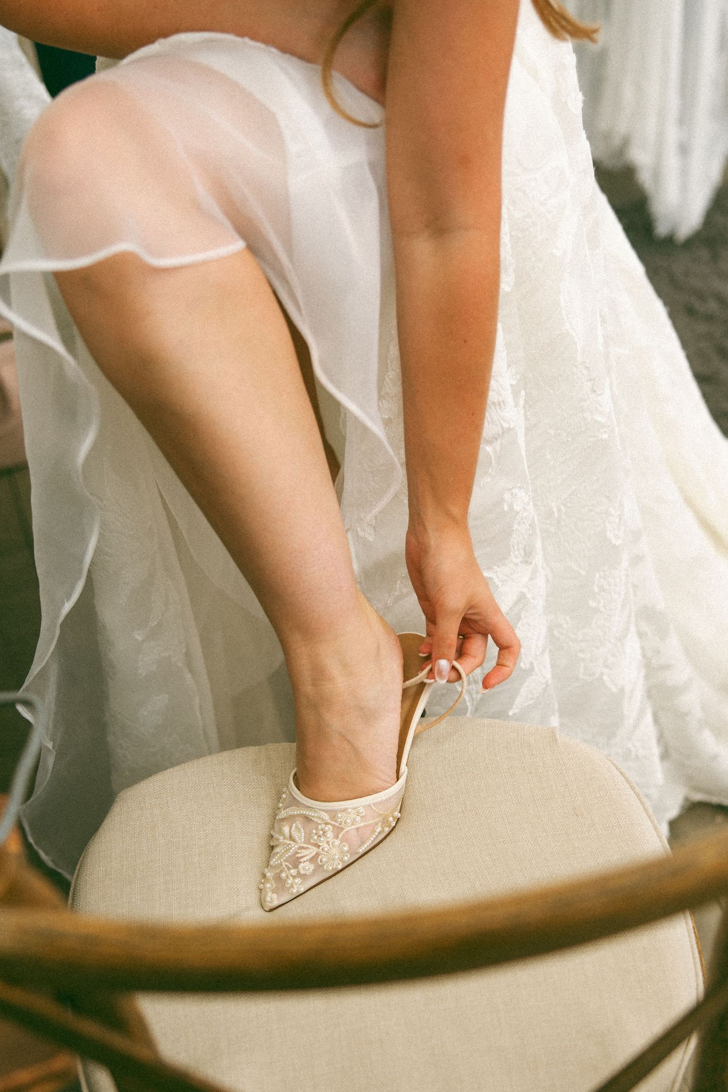 Bride in a lace wedding gown with sheer tulle skirt fastening the strap of an embroidered pointed bridal heel on a cushioned chair.