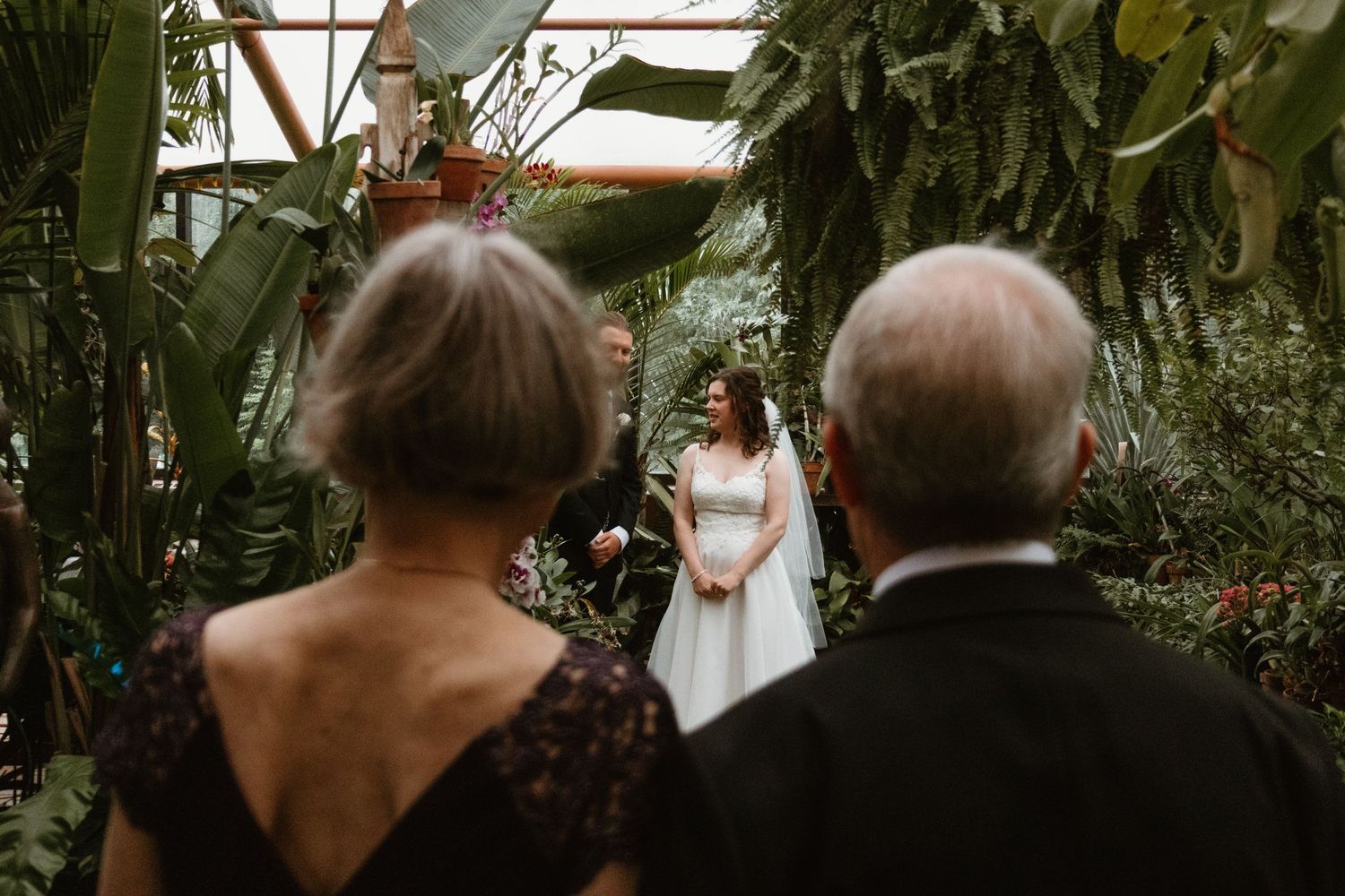 Two wedding guests watch a bride in a white gown stand at the altar surrounded by lush tropical plants in a greenhouse venue.