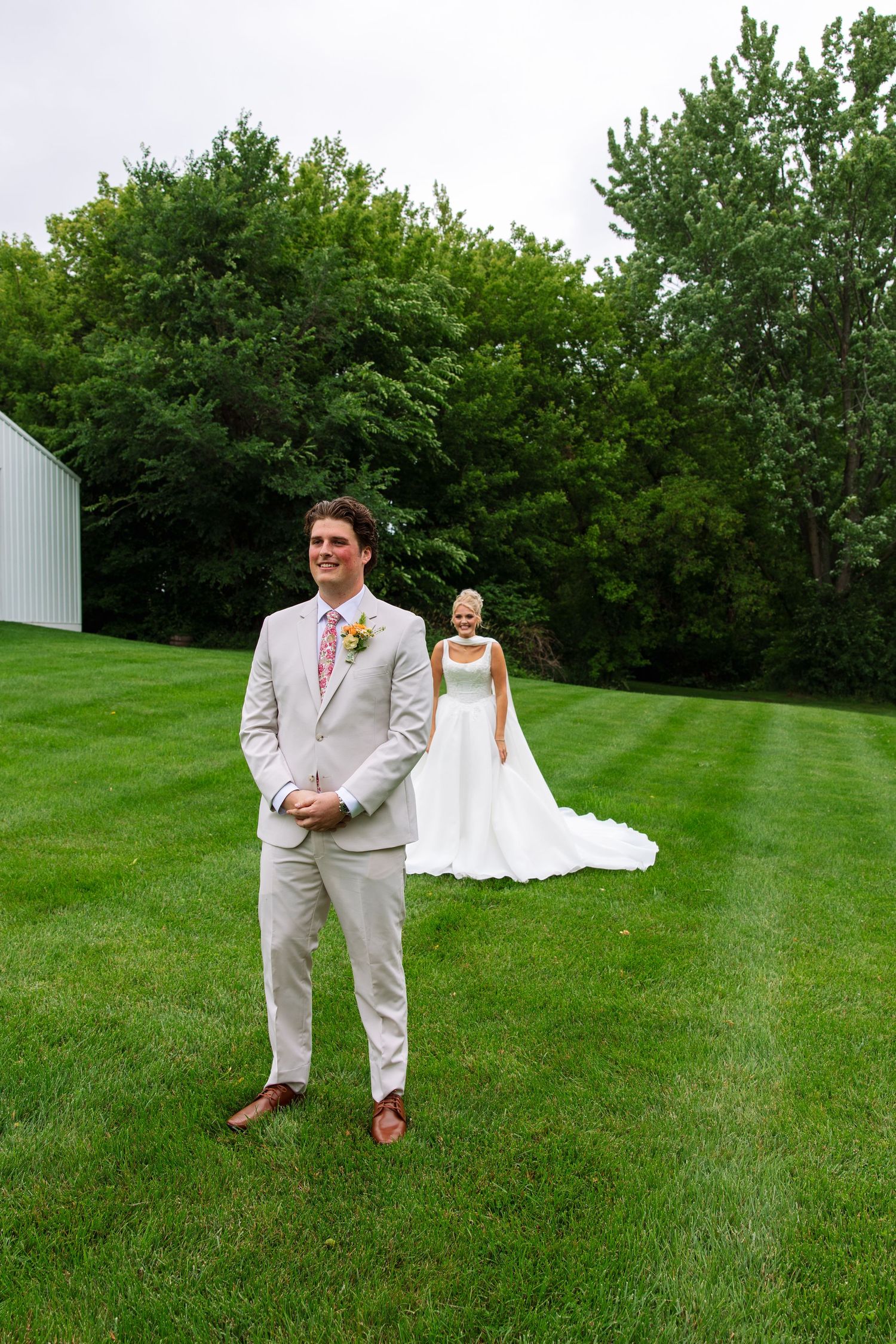 Groom in a tan suit smiles with hands clasped during a first look as the bride in a white gown approaches on a green lawn.