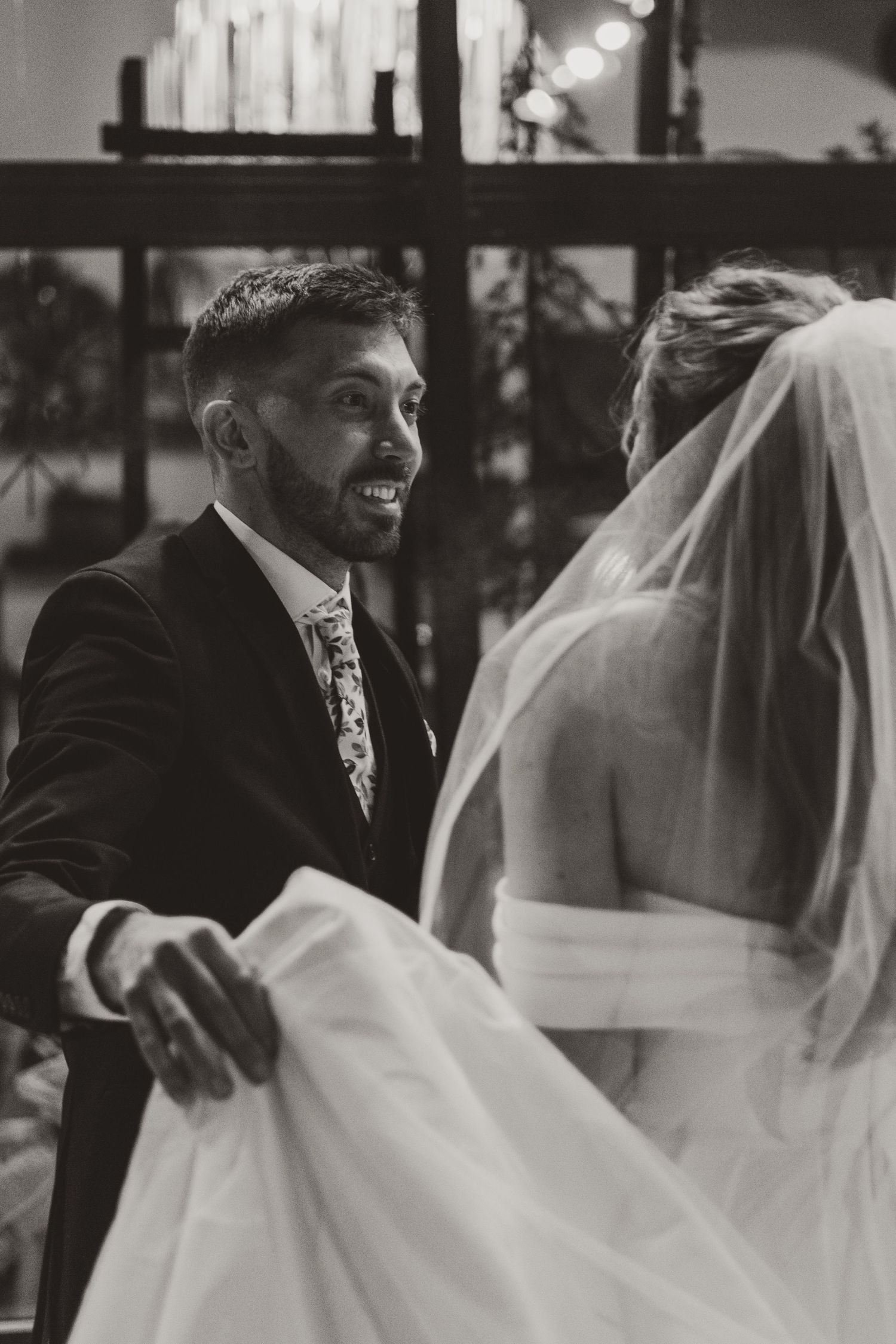Smiling groom in a dark suit and patterned tie holds the bride's gown skirt as she faces him in her veil.