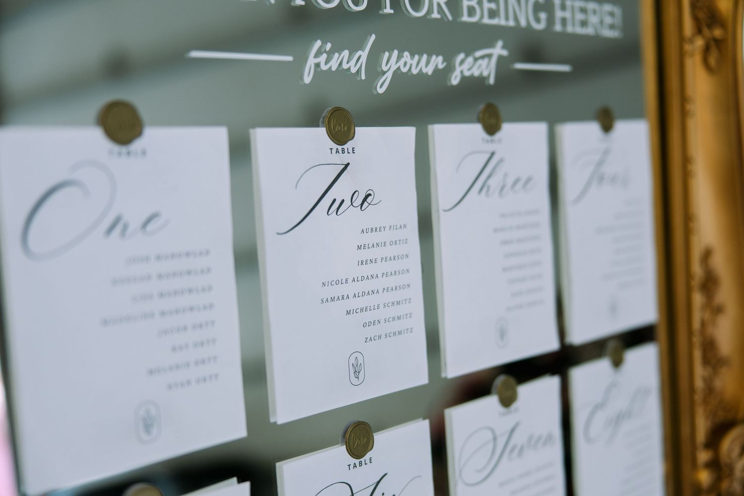 Close-up of elegant wedding seating chart cards with calligraphy table names pinned to a mirror in an ornate gold frame.