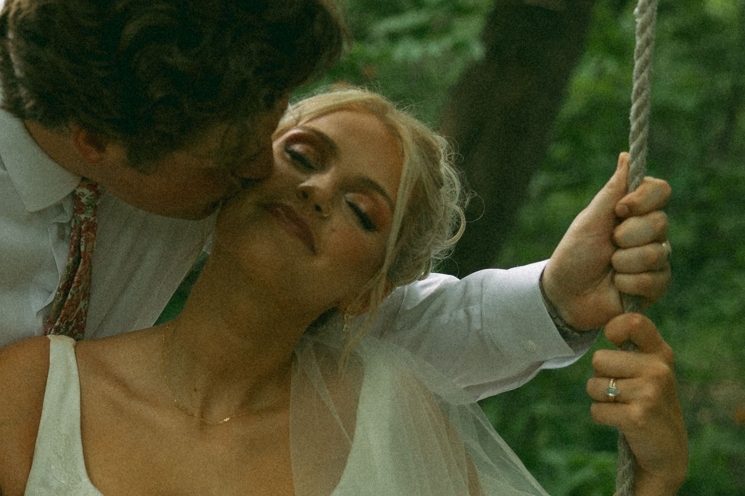 Groom kisses the smiling bride on the cheek as she holds a rope swing in a wooded outdoor wedding portrait.