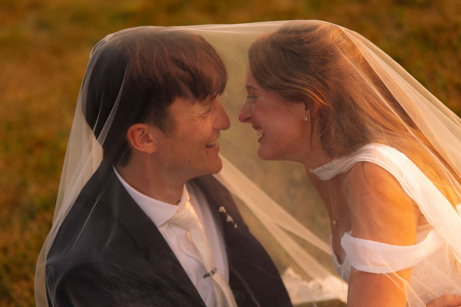 Smiling bride and groom share a joyful moment beneath a sheer veil in warm golden sunset light at their wedding.