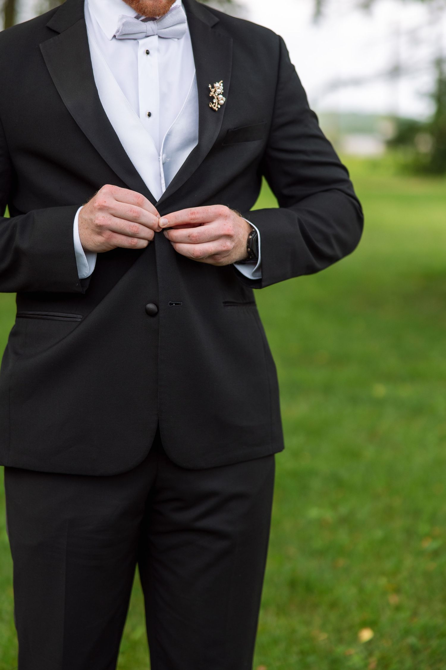 Groom in a black tuxedo with a gray bow tie and floral boutonniere buttons his jacket while standing on a green lawn outdoors.