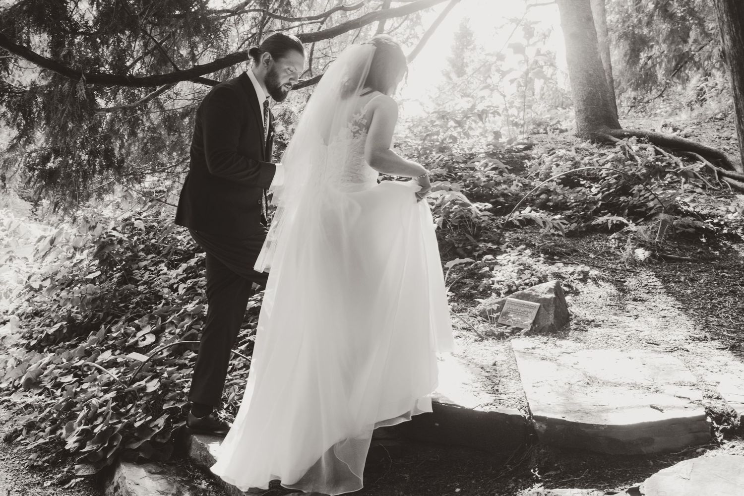 Black and white photo of a bride in a flowing gown and veil stepping onto stone slabs beside a groom in a suit in a wooded outdoor setting.