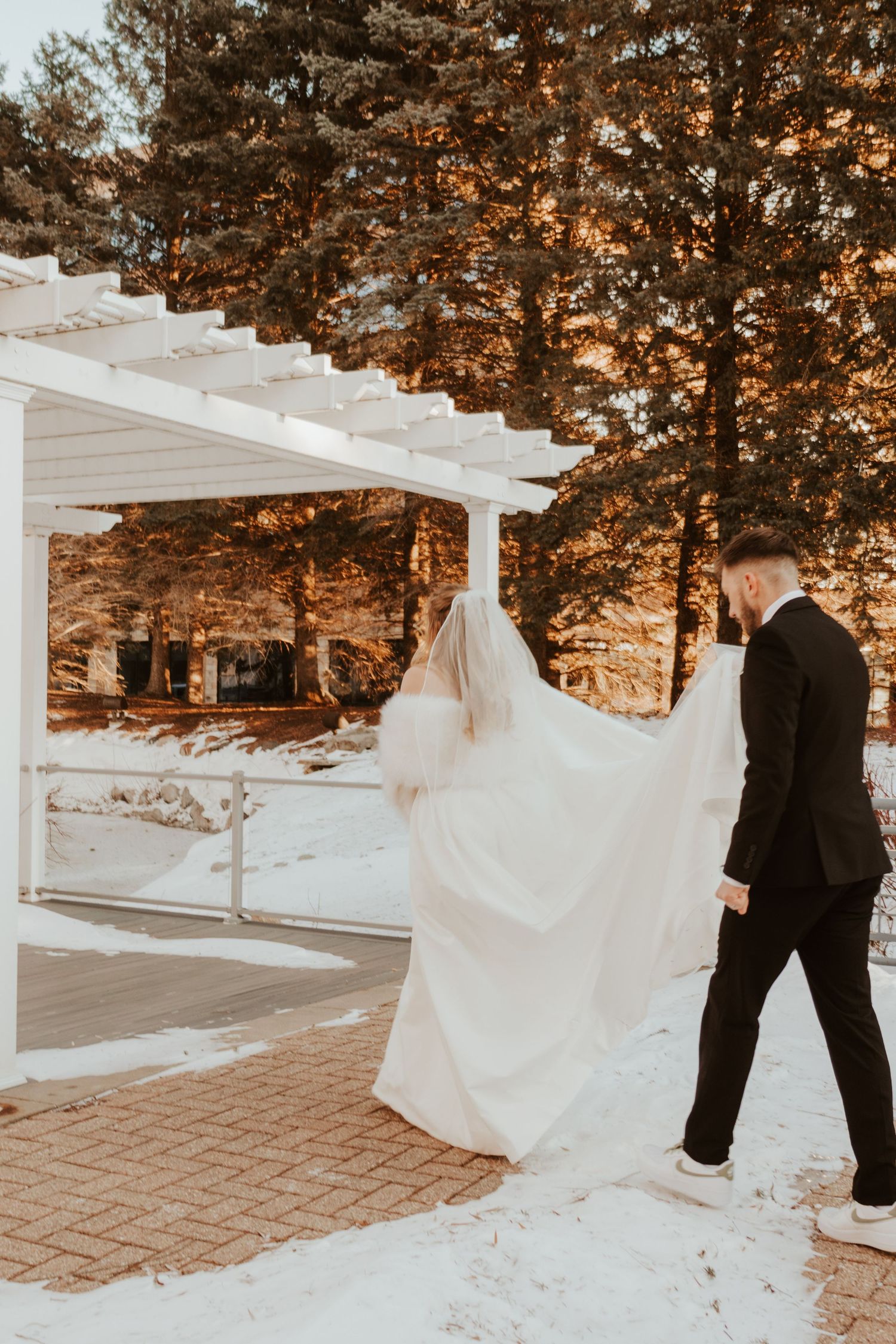 Bride in a white gown with a long veil and groom in a black suit walk through snow beneath a white pergola.