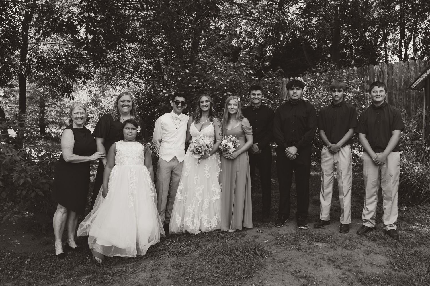 Black and white group portrait of a wedding party standing together outdoors, including a flower girl in a white dress.