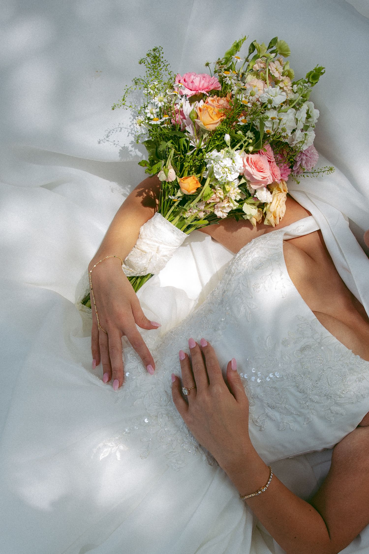 Bride in a lace wedding dress holds a colorful bouquet of pink, peach and white flowers over her face.