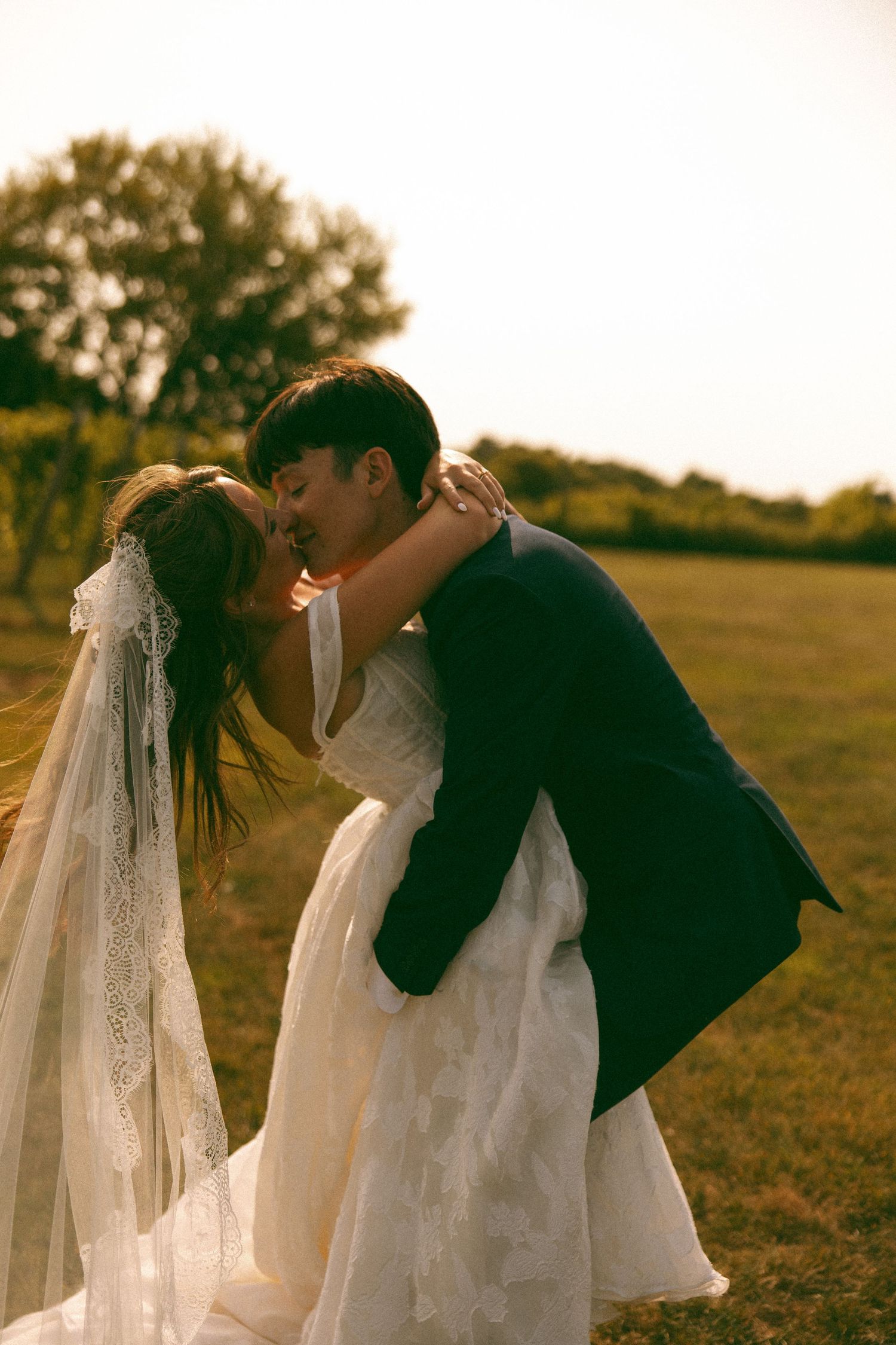 Groom dips and kisses his bride in a lace gown and veil on a sunlit grassy field at golden hour.