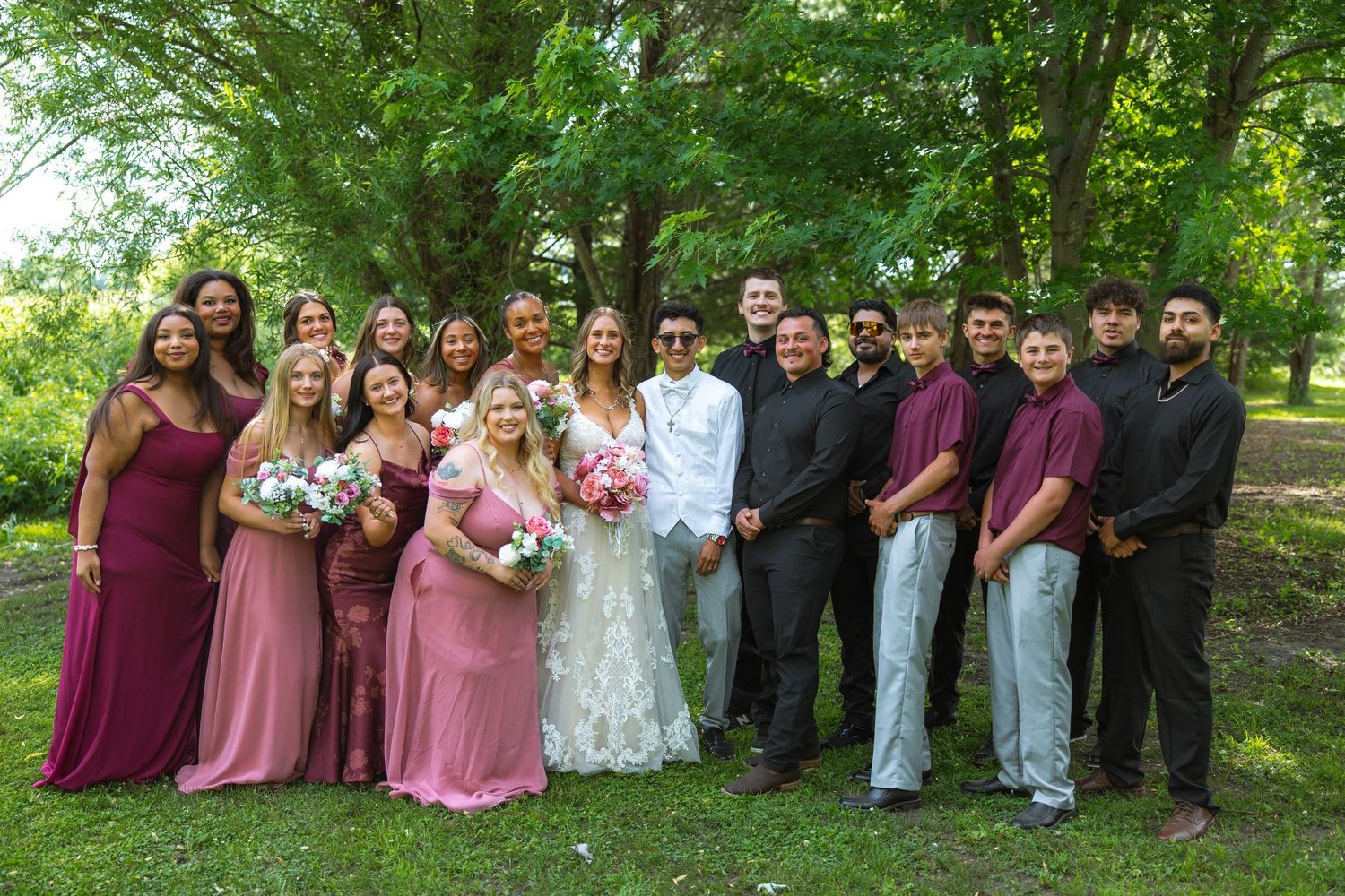 Bride and groom pose with bridesmaids in mauve dresses and groomsmen in dark attire on a sunny green lawn under leafy trees.