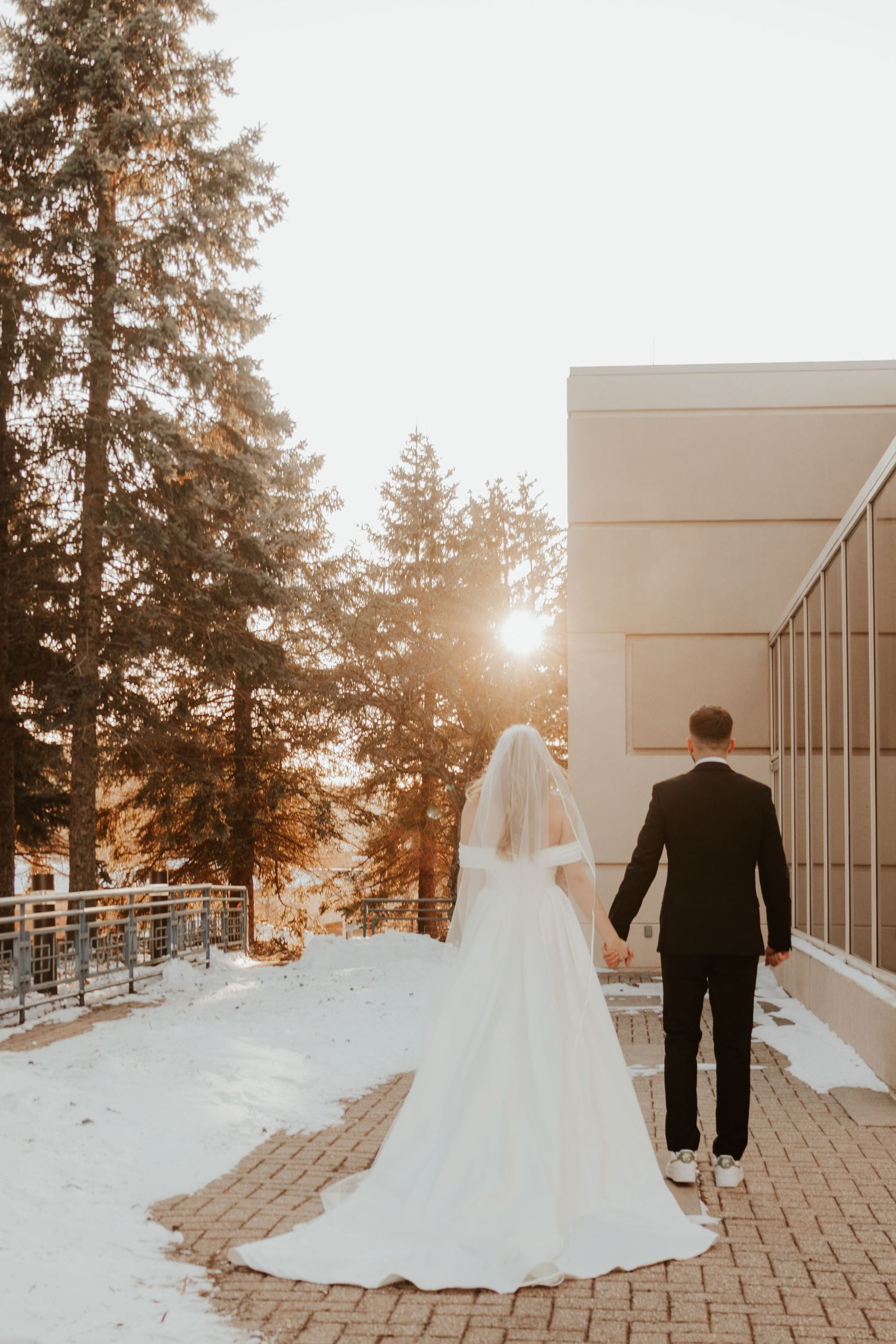 Bride and groom hold hands and walk away on a brick path through snow, with golden sunlight filtering through evergreen trees.