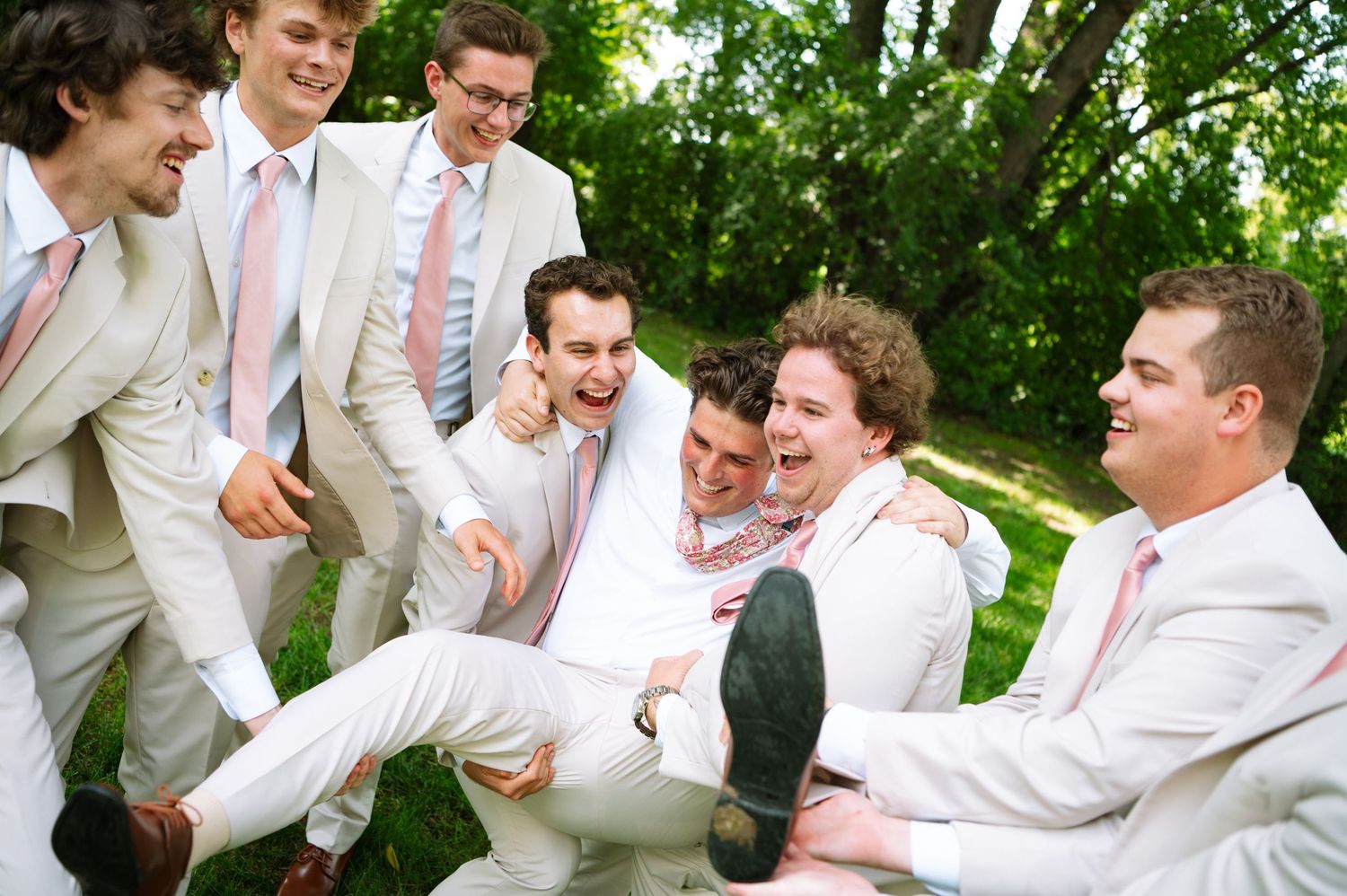 Laughing groomsmen in light suits and pink ties playfully lift the groom while celebrating outdoors among green trees.