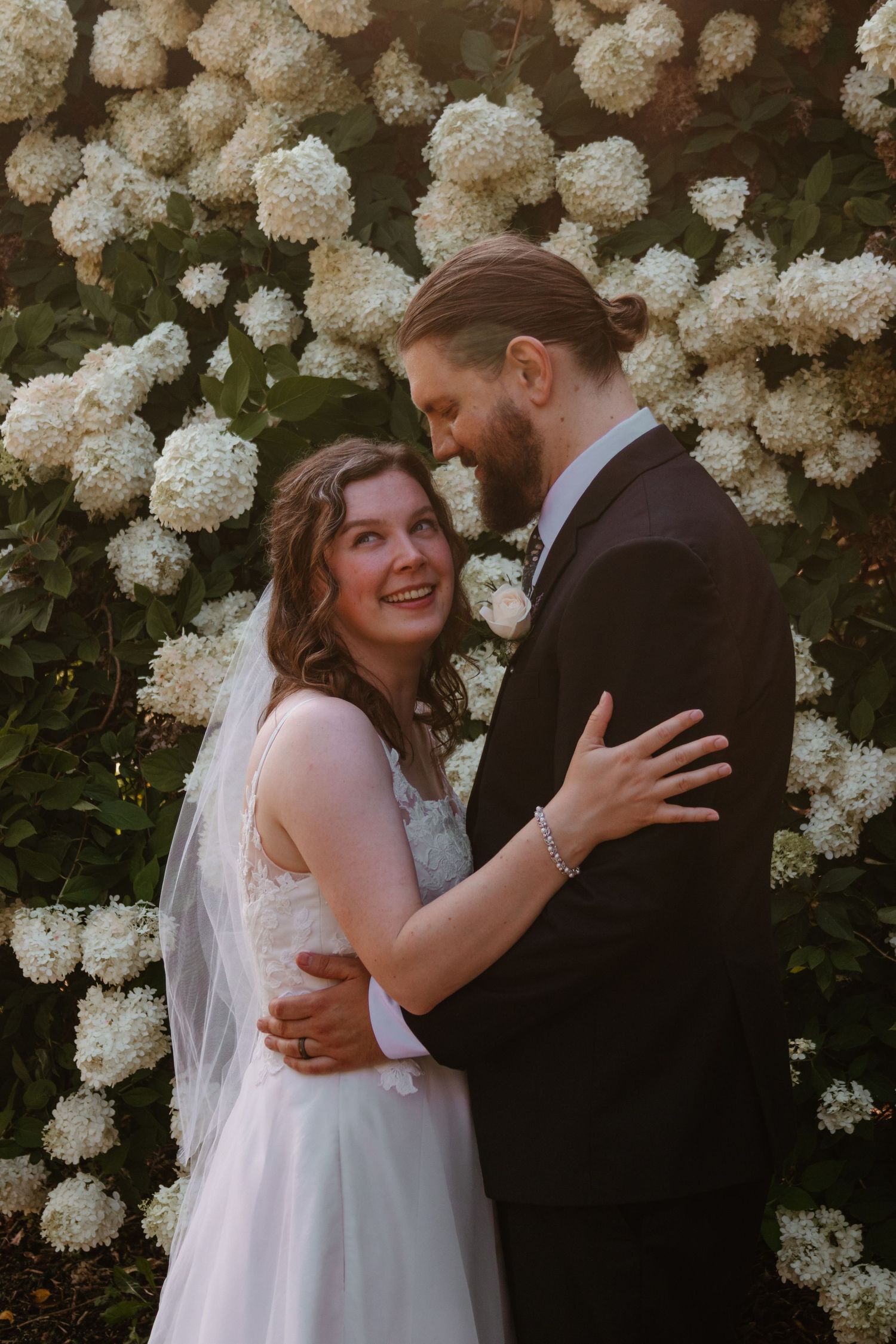 Smiling bride in a lace gown and veil embraces her bearded groom in a dark suit in front of blooming white hydrangea bushes.
