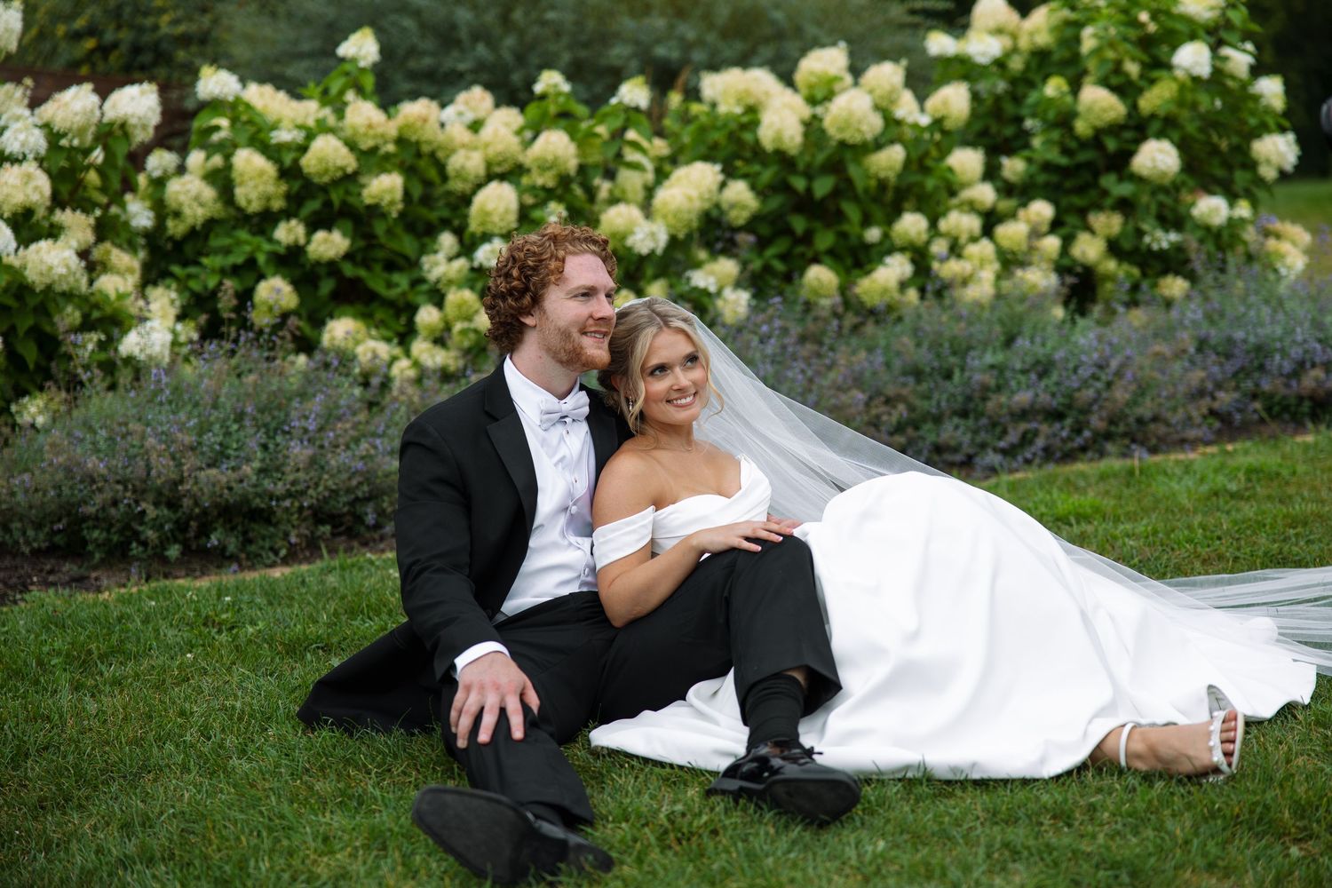 Smiling groom in a black suit sits close to his bride in a white gown on the grass beside blooming white hydrangeas.