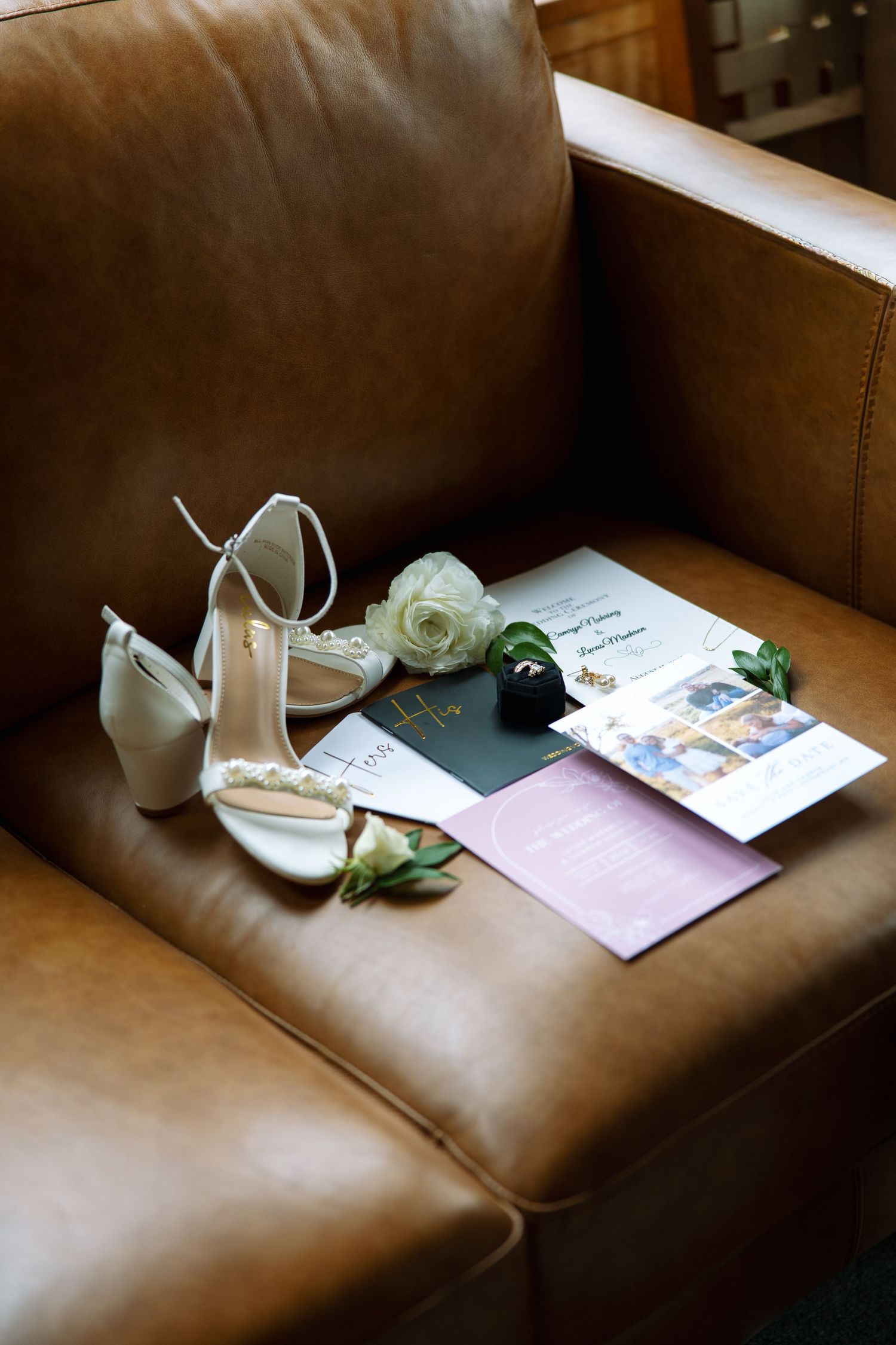 Cream strappy wedding heels, white rose boutonniere, and invitation suite resting on a tan leather sofa.