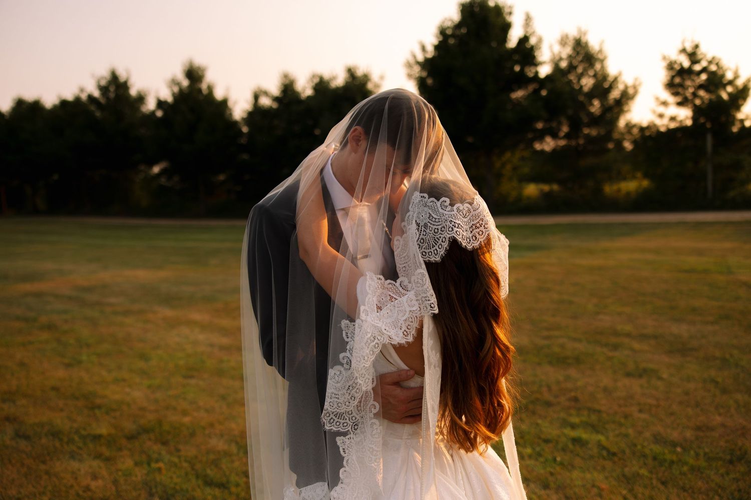 A bride and groom kiss beneath a lace-trimmed wedding veil in a grassy field at golden hour sunset.