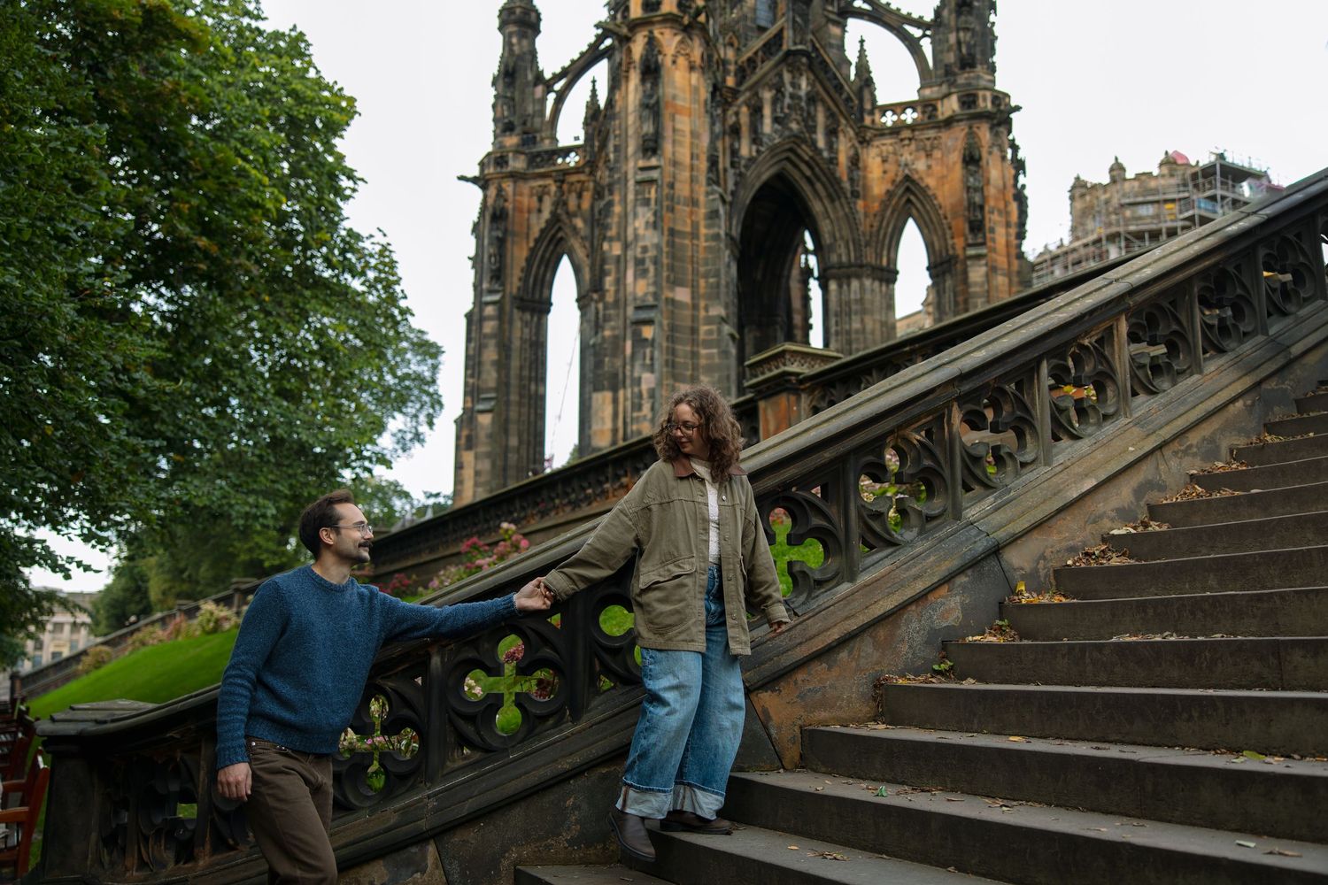 Couple holding hands descending stone steps beside a gothic monument during an engagement photo session.