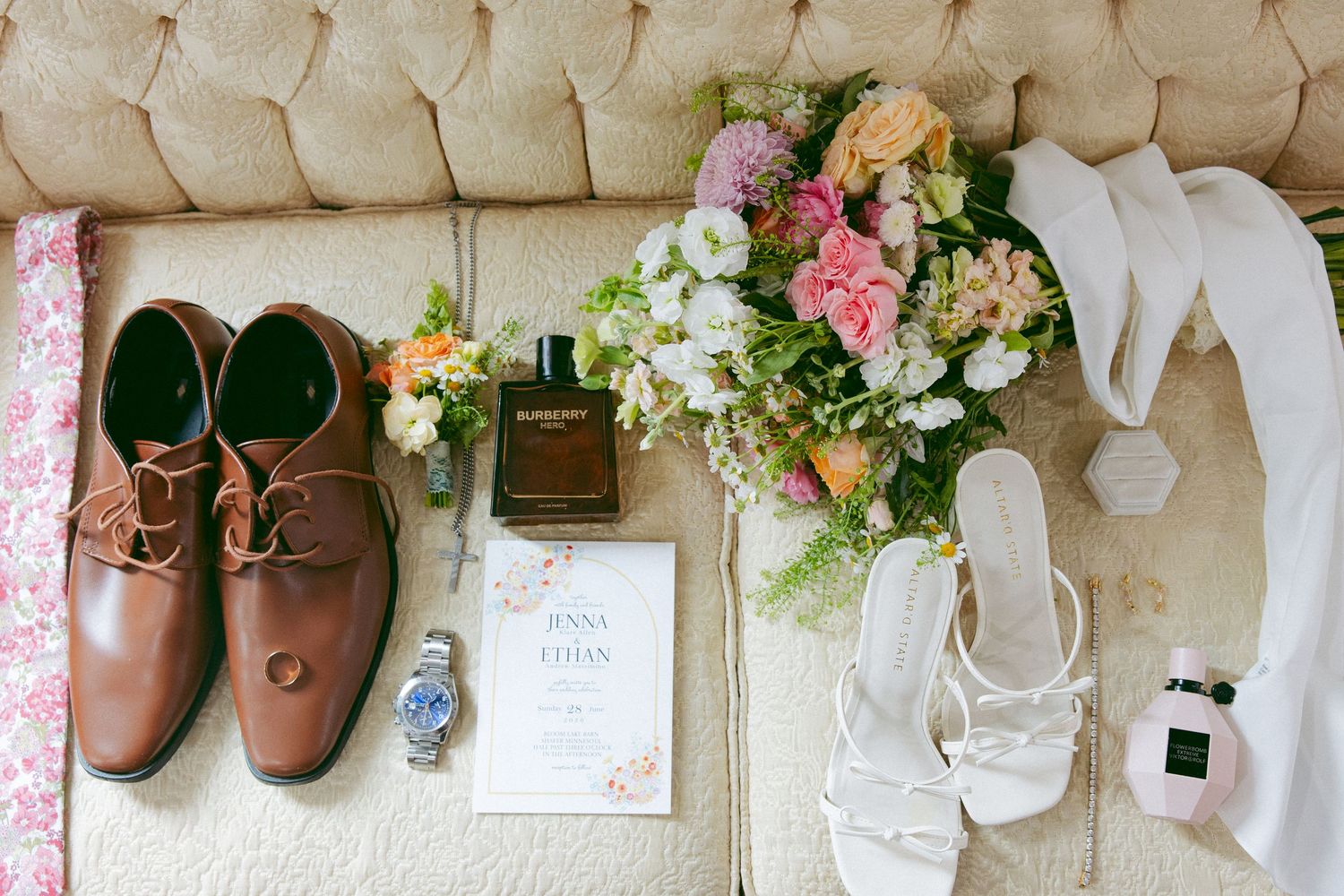 Wedding flat lay with brown leather groom shoes, white bridal heels, a pastel floral bouquet, cologne, watch, ring and invitation on a tufte