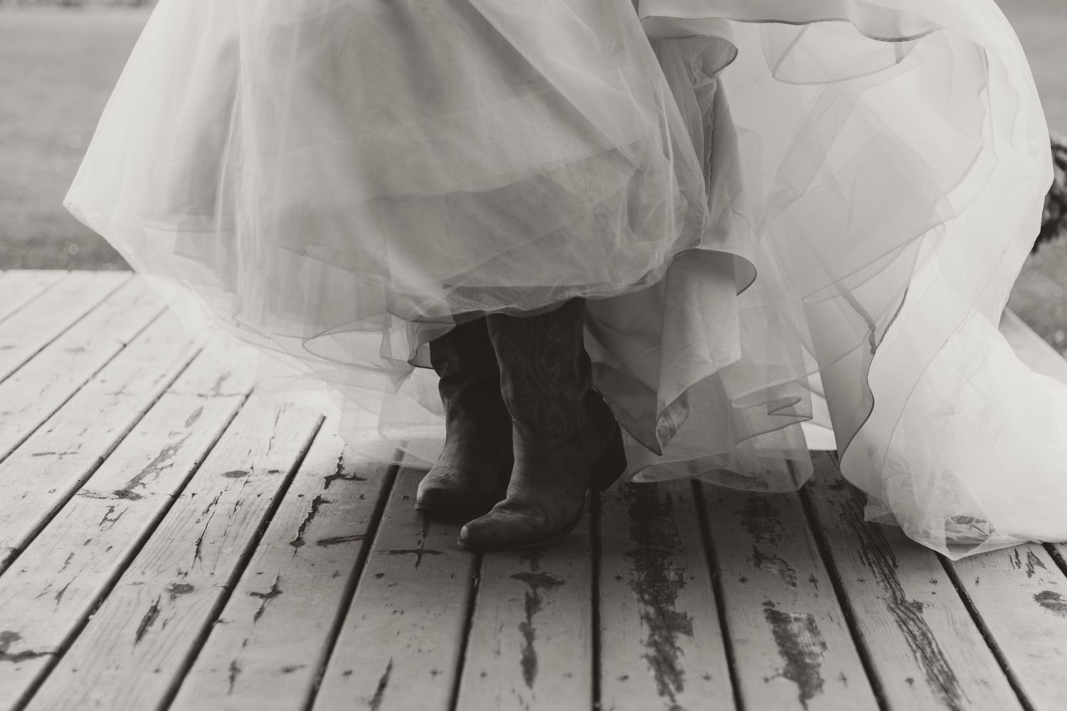 Black and white photo of a bride in a flowing tulle gown wearing rain boots while standing on a weathered wooden deck.