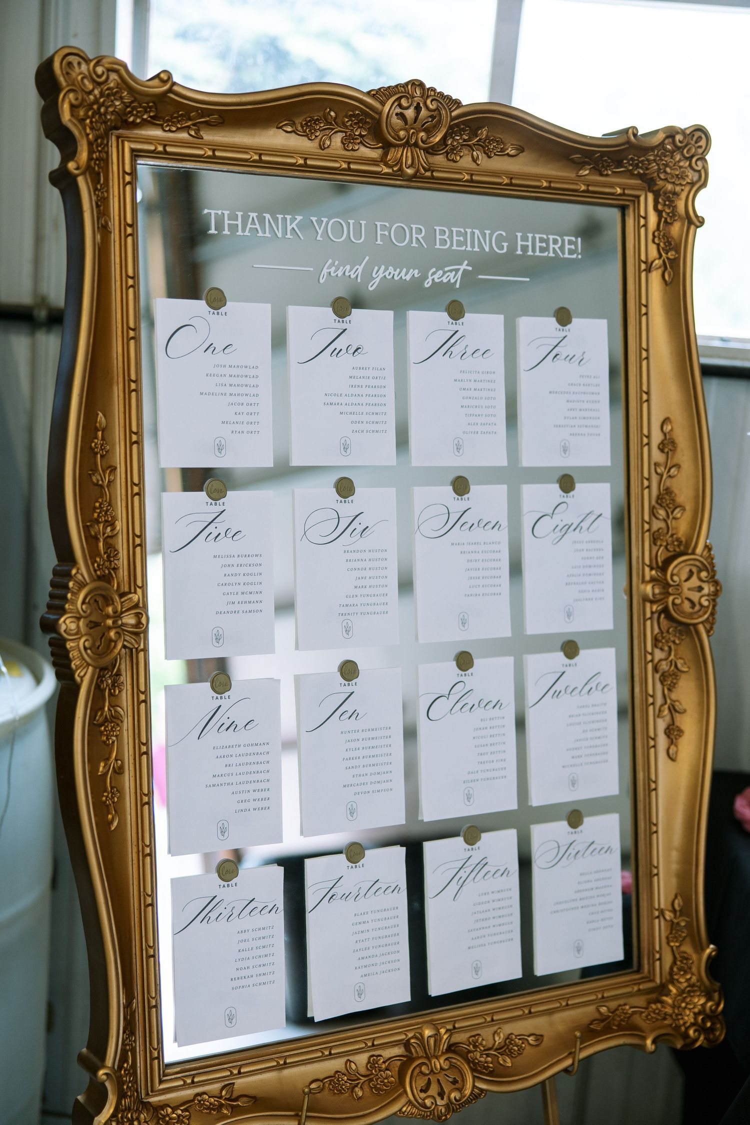 Wedding seating chart with calligraphy cards for tables one through fifteen pinned to a mirror in a gold baroque frame.