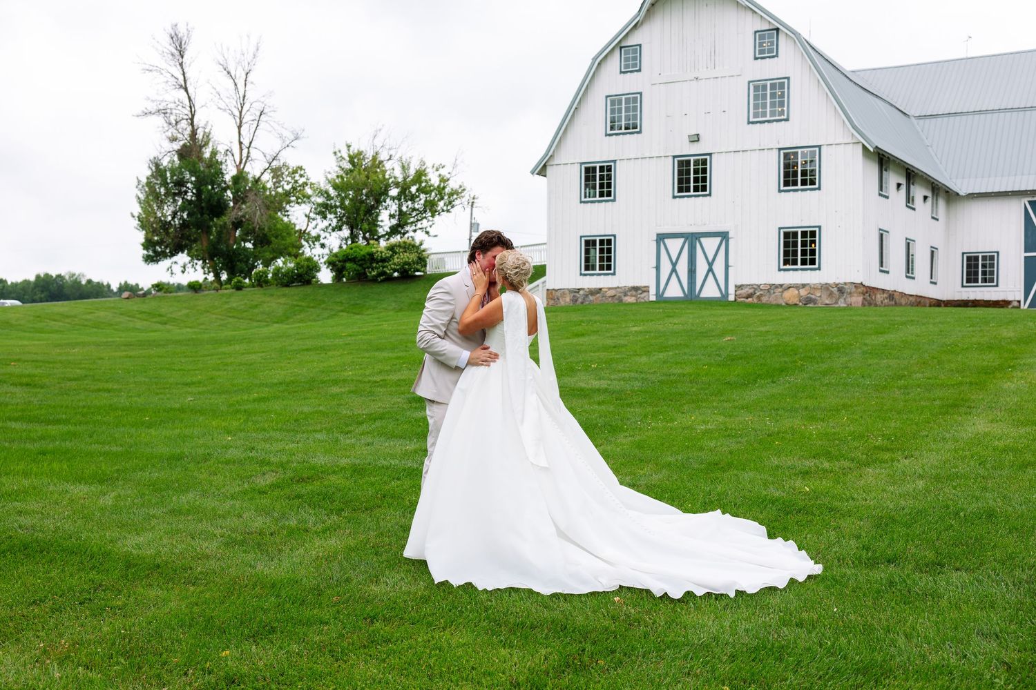 Bride and groom embrace on a lush green lawn in front of a white barn wedding venue under a soft cloudy sky.