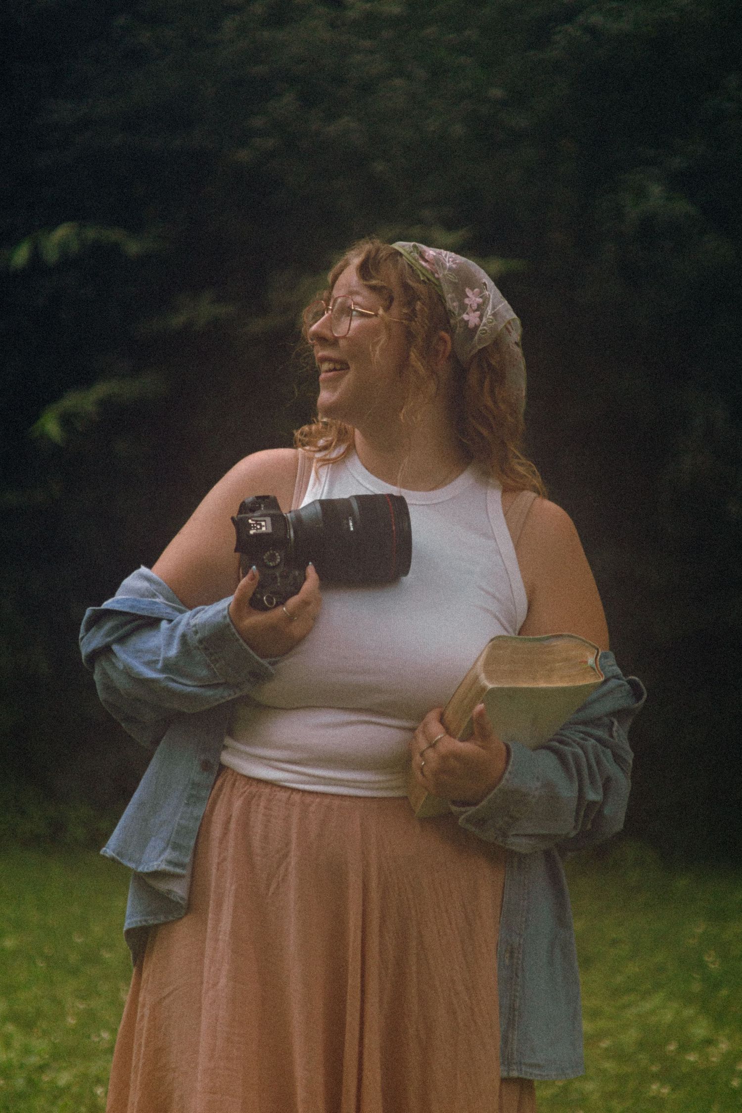 Smiling woman in a floral headscarf, white tank top and peach skirt holds a camera and a book while standing on grass near trees.