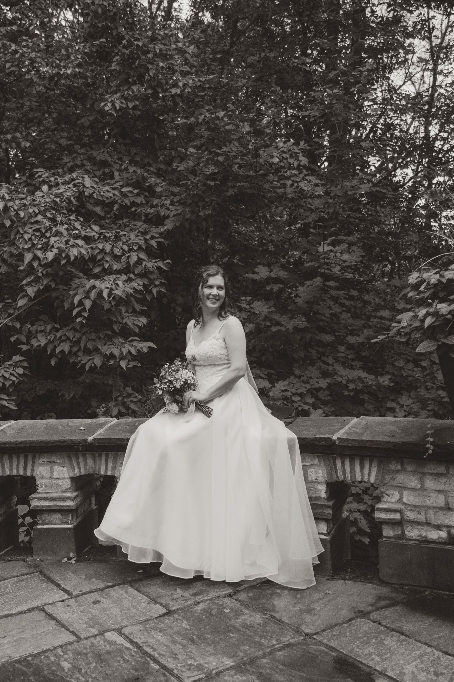 Black and white portrait of a bride in a flowing gown holding a bouquet while seated on a stone wall in front of lush trees.