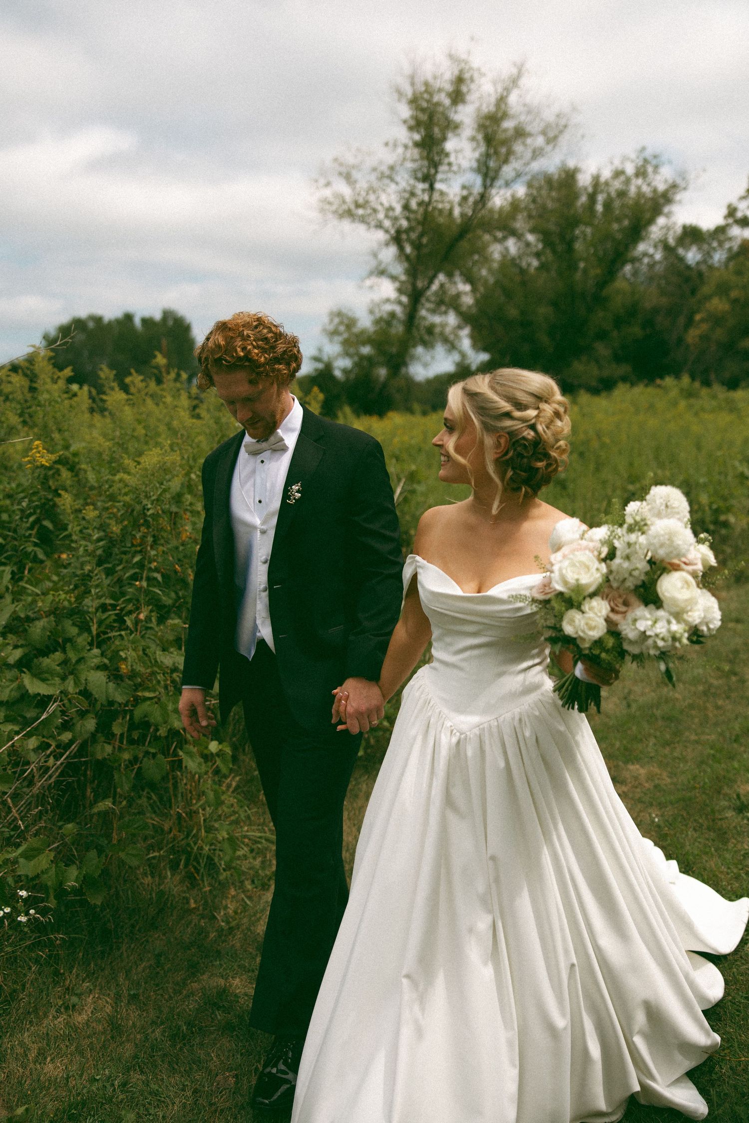 Bride in a strapless satin wedding gown holding a white bouquet walks hand in hand with a groom in a black suit through a sunny field.