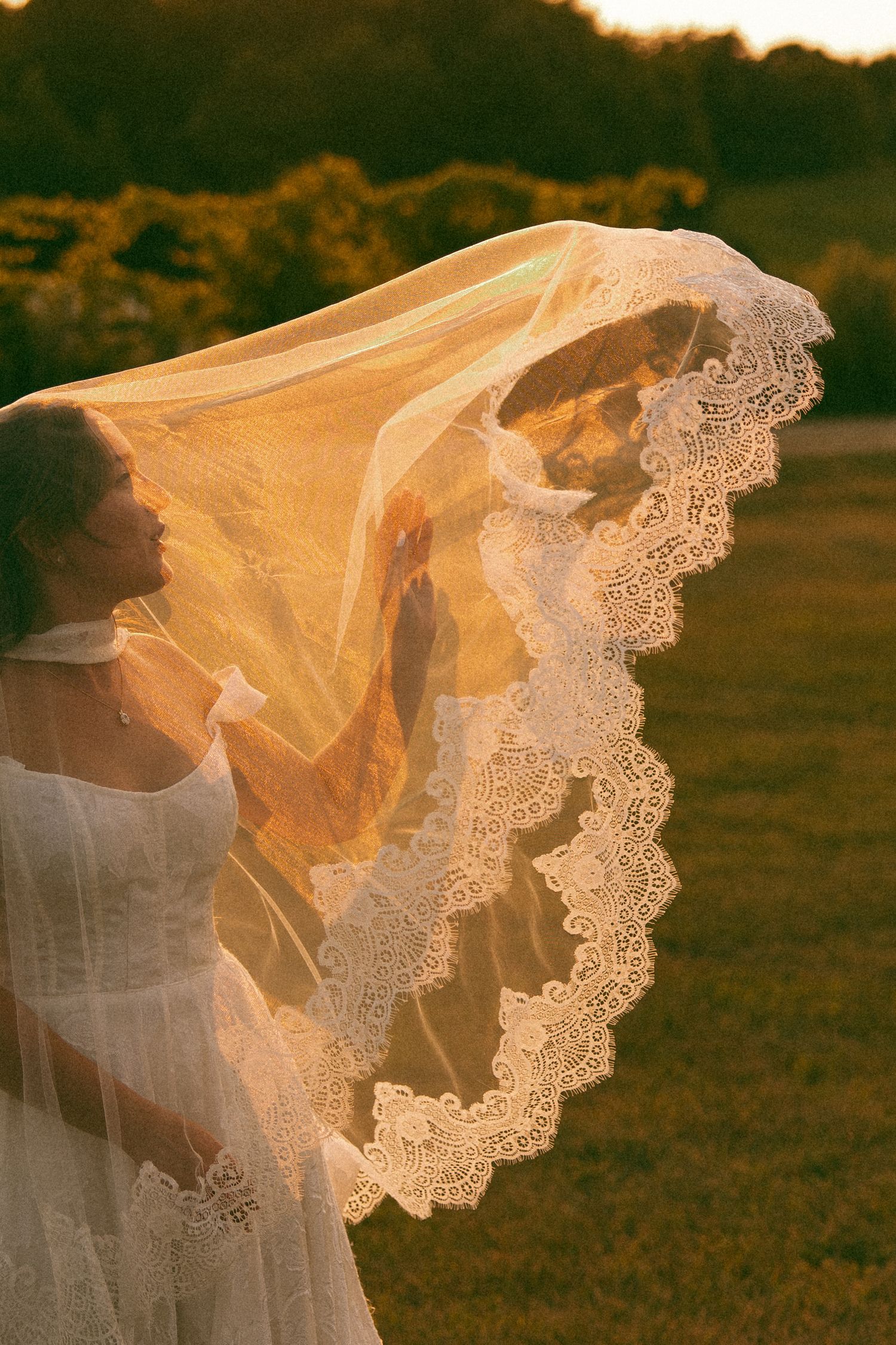 Bride in a white dress holds up a lace-trimmed veil that billows in the breeze, backlit by warm golden sunset light.