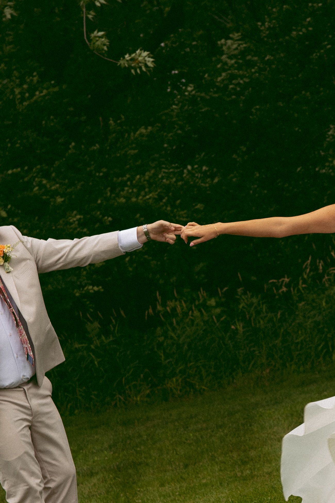 A groom in a beige suit with a floral boutonniere reaches out to hold hands with a bride outdoors on a grassy lawn.
