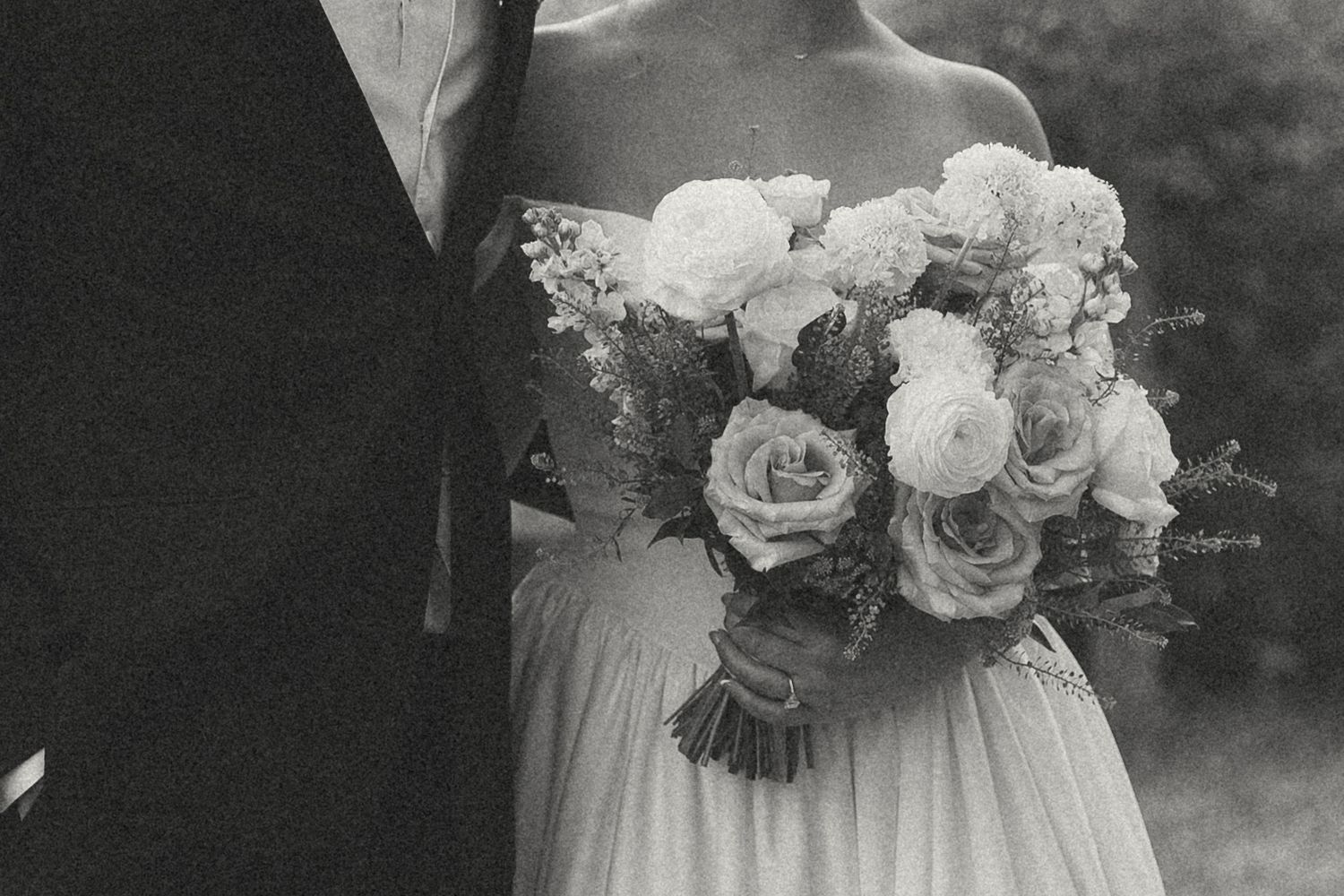 Black and white photo of a bride in a strapless gown holding a romantic bouquet of roses beside a groom in a dark suit.