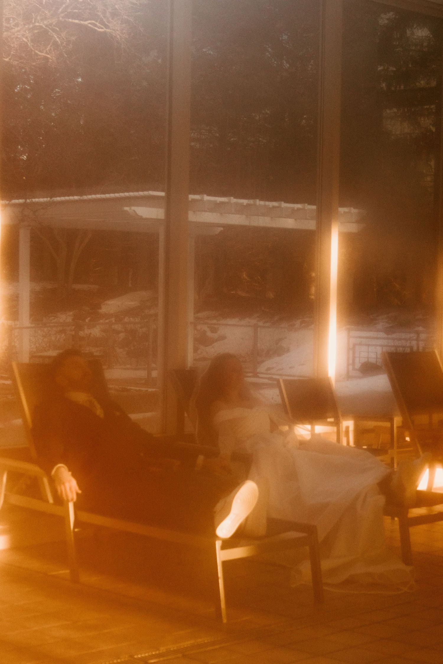 Newlyweds relax on lounge chairs by large glass windows with a warm golden light flare and a snowy view outside.