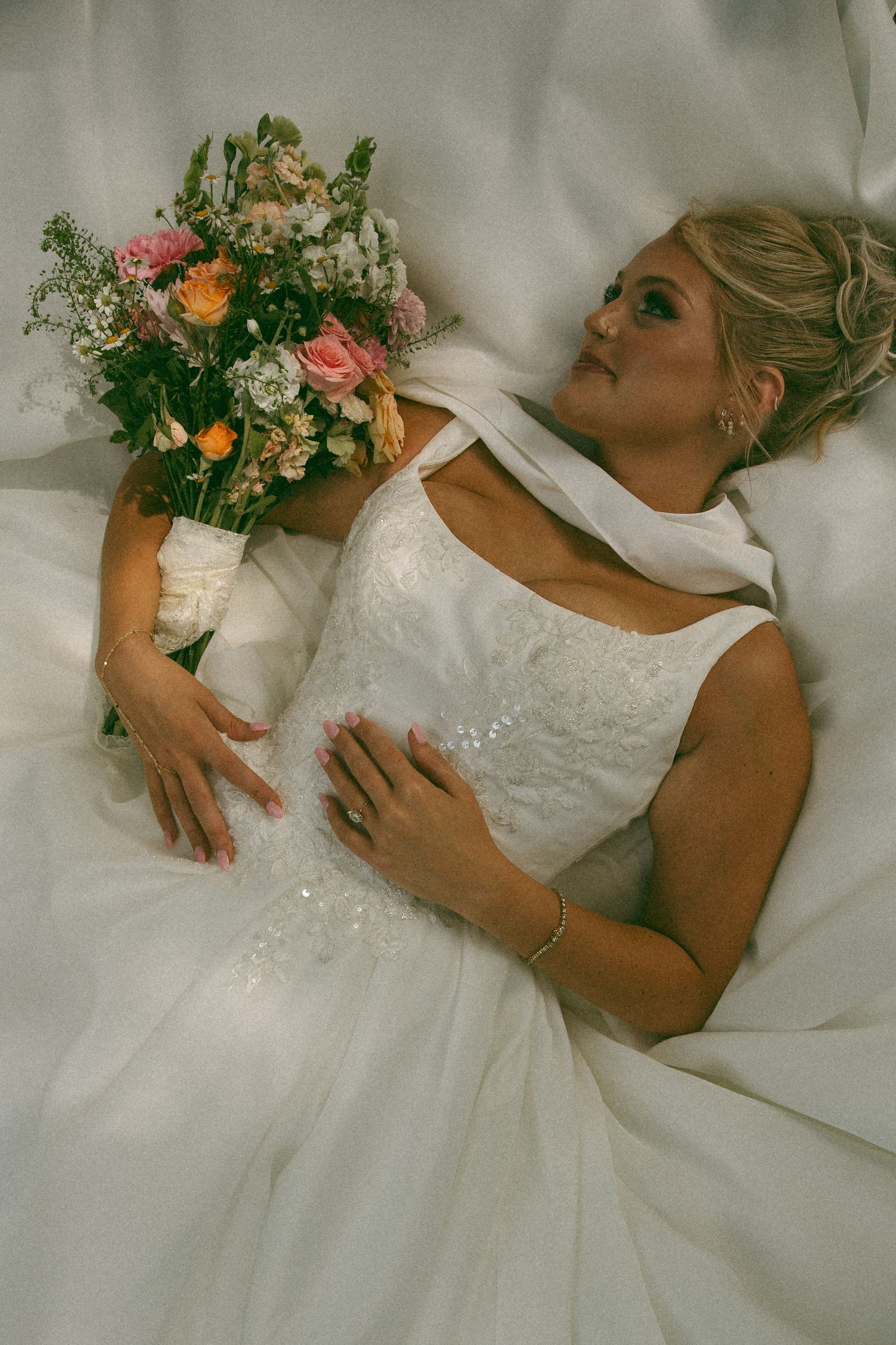 A bride in a white wedding gown lies back holding a bouquet of pink, orange, and white flowers, gazing upward.