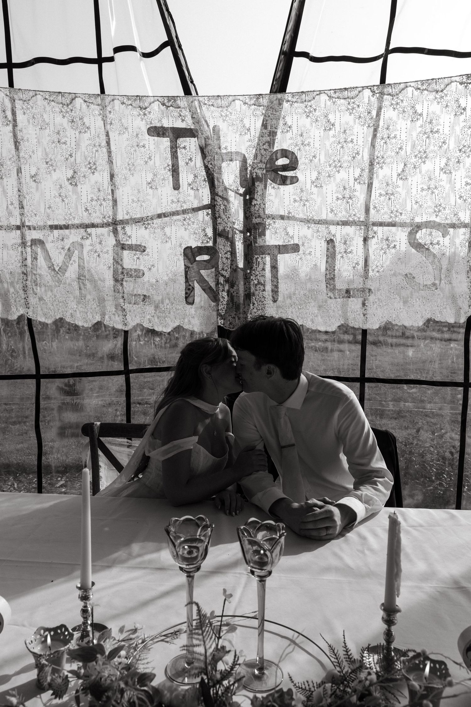 Black and white photo of a newlywed couple kissing at a reception table beneath a lace banner and candlelit centerpieces.