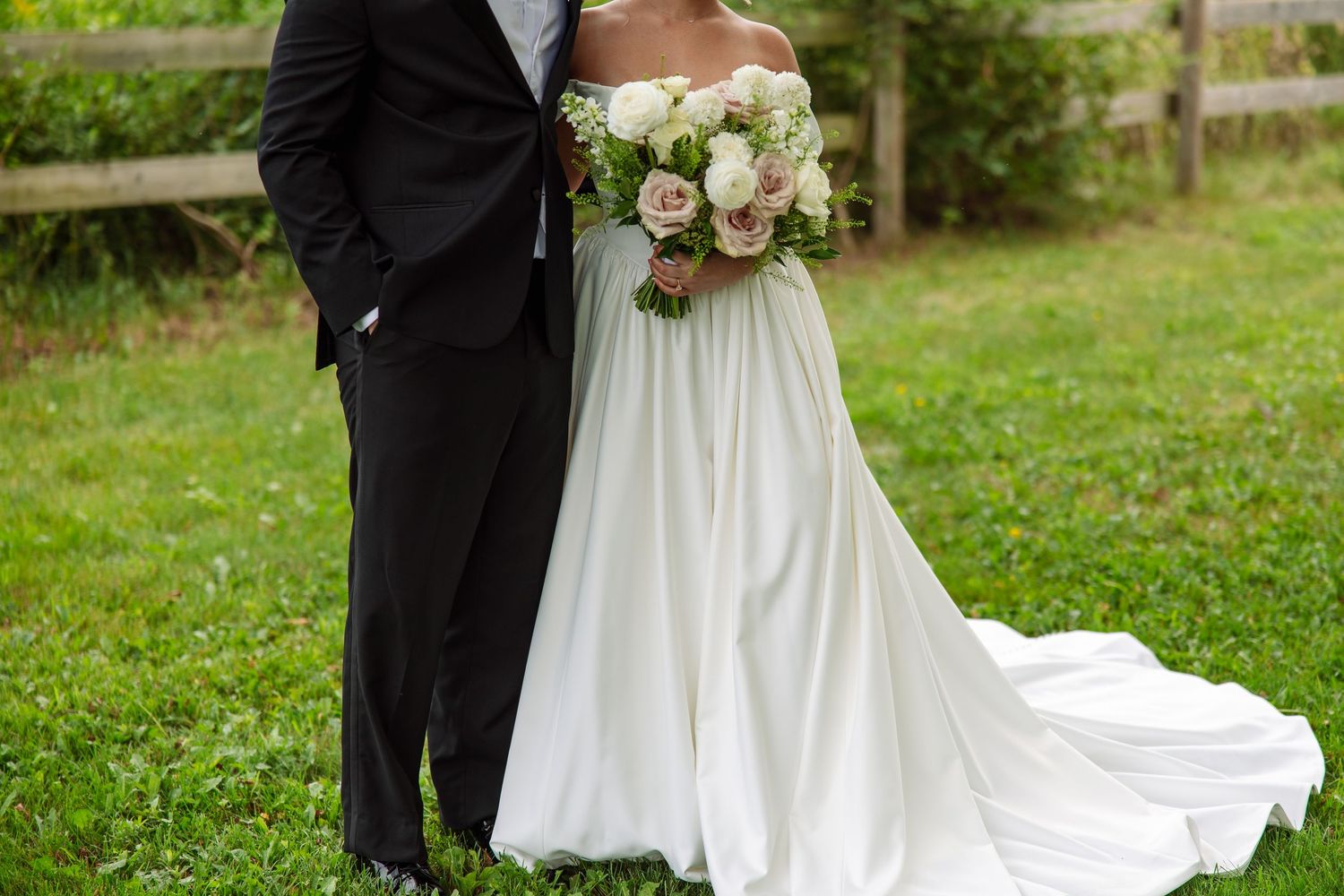 Bride in a flowing white satin gown holding a white and blush rose bouquet stands beside a groom in a black suit on a green lawn.
