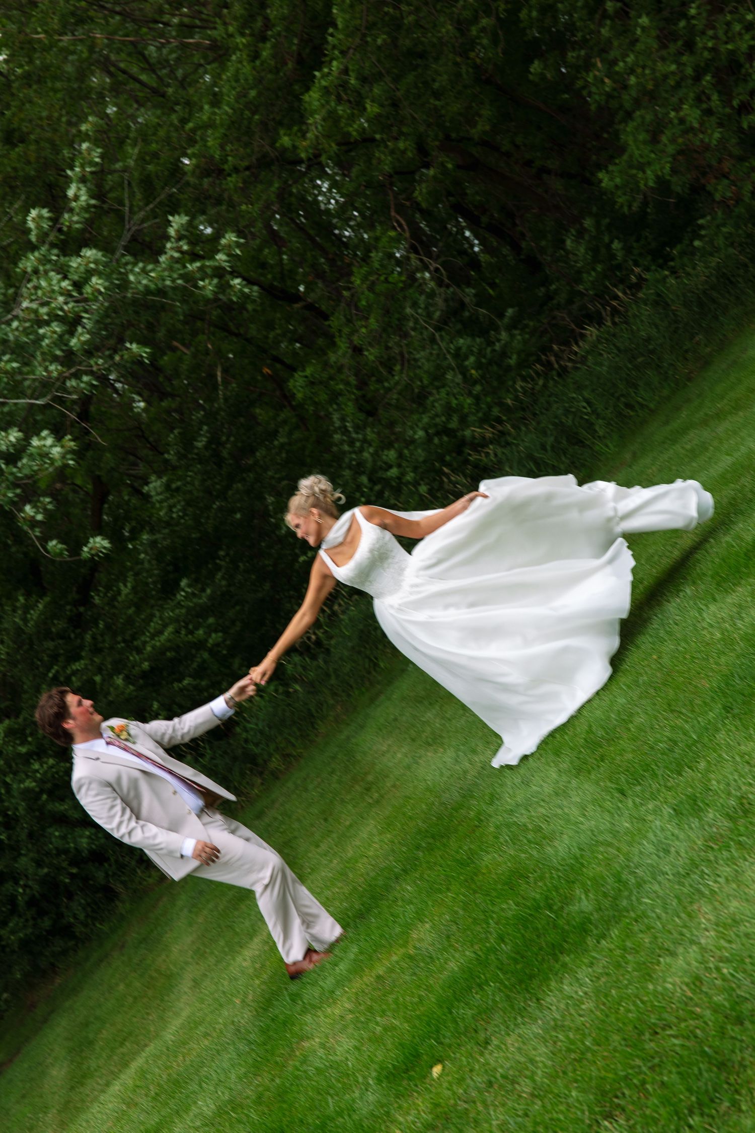 Bride in a flowing white gown holds hands with the groom in a light suit while leaning playfully on a green grassy hillside.