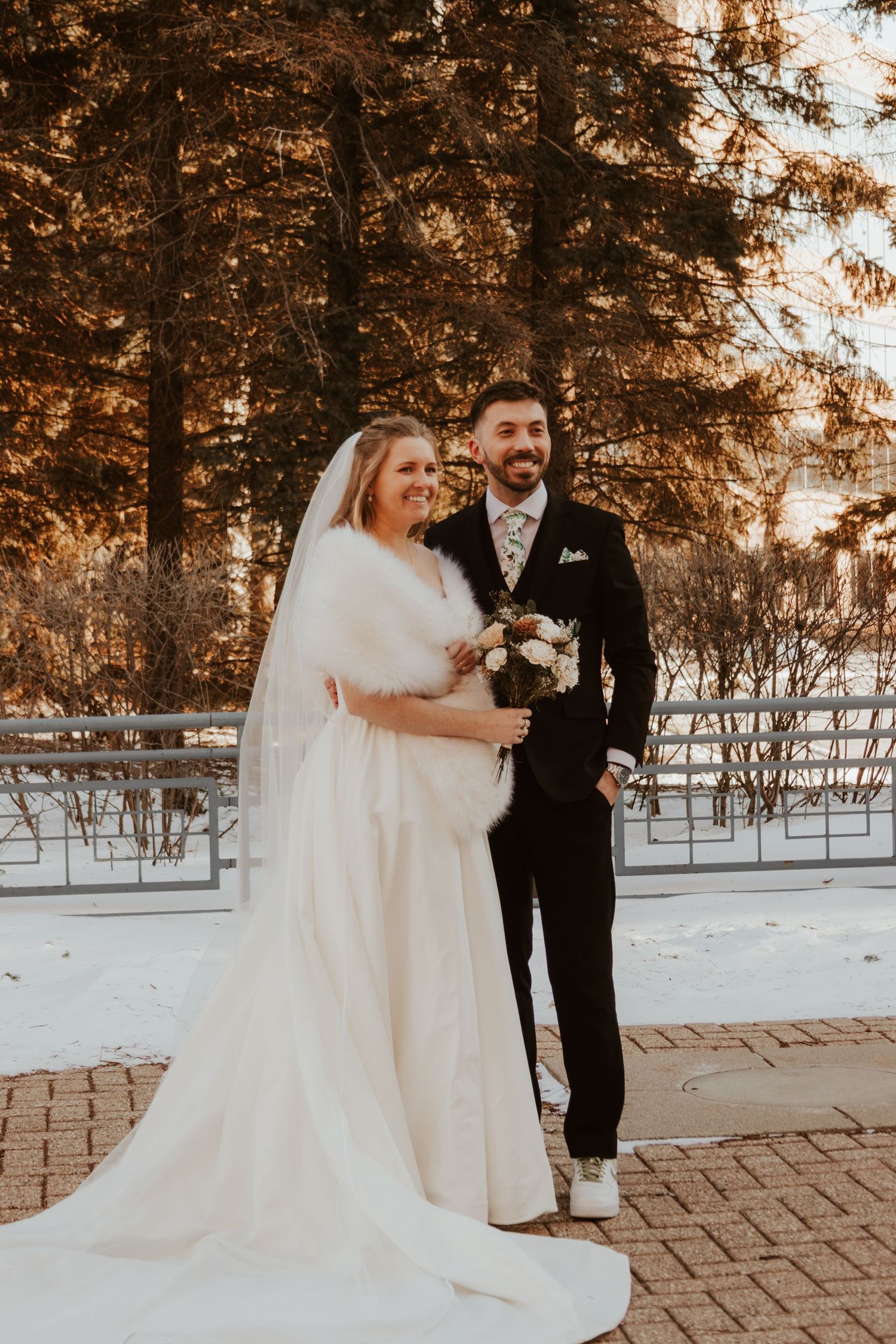Smiling bride in a fur wrap and long veil poses with her groom in a black suit among snowy evergreen trees.