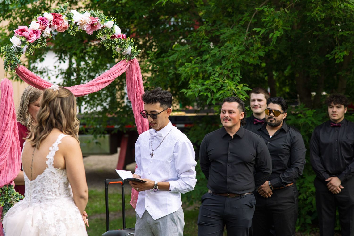 Officiant reads from a paper during an outdoor wedding ceremony while the bride faces him and groomsmen in black look on.