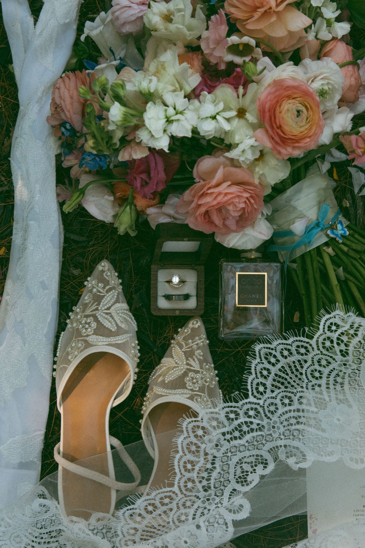 Bridal flat lay with peach and pink bouquet, lace-embellished heels, ring box, perfume bottle, and lace veil on grass.