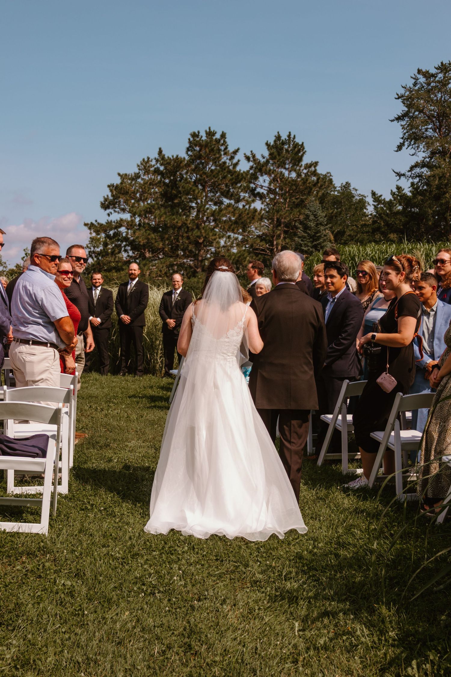 Bride in a long veil and white gown walks down the grass aisle at an outdoor wedding as guests stand and watch.