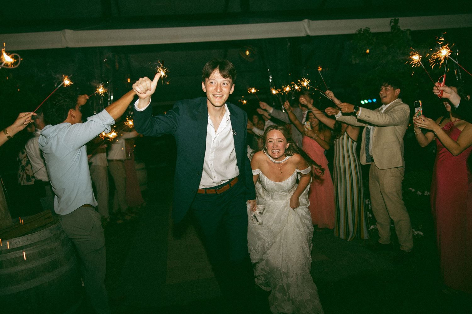 Smiling groom and bride walk through a sparkler send-off lined by cheering wedding guests holding lit sparklers at a nighttime reception.