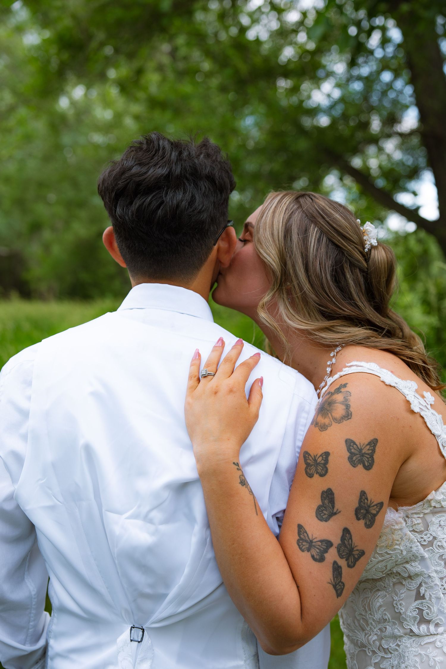 Bride with butterfly tattoos on her arm kisses the groom on the cheek, with green trees softly blurred in the background.