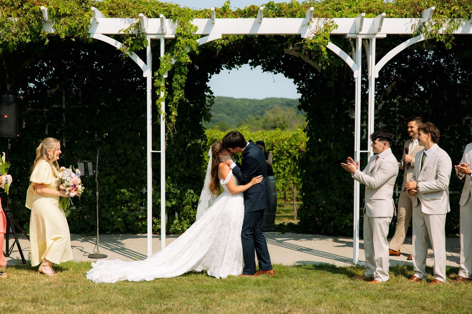 A bride and groom share their first kiss under a white pergola as groomsmen applaud and guests cheer at an outdoor wedding.